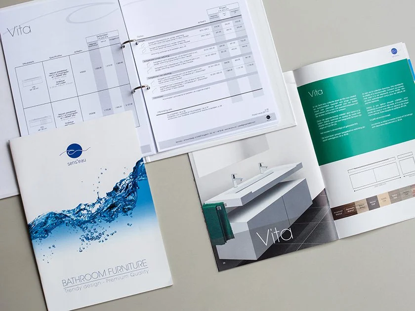 Open bathroom furniture catalog with the title "Vita" and a brochure showing a modern double sink vanity, along with a closed sample booklet titled "sens'eau" featuring water splash imagery and the description "Bathroom Furniture, Trendy design, Prem