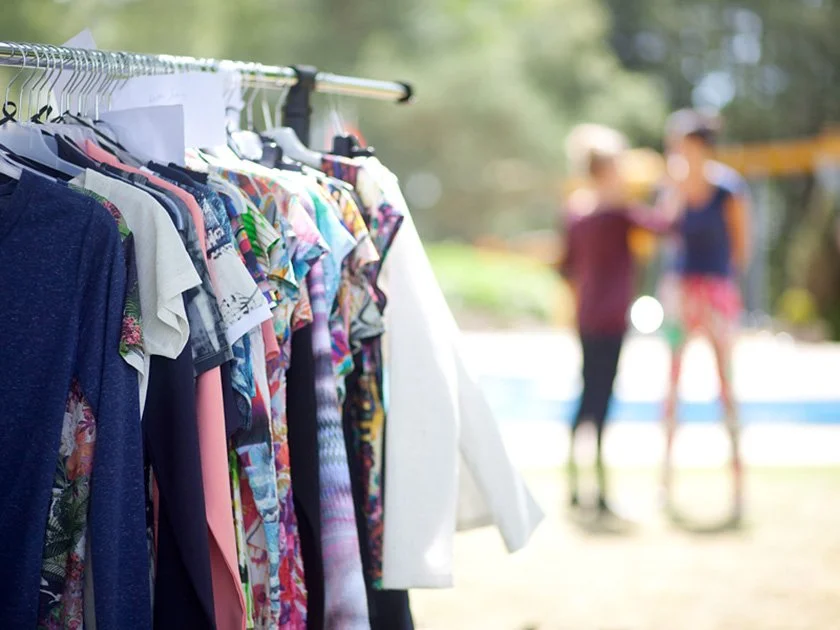 Clothes on a rack, with two people blurred in the background at an outdoor yard sale or flea market.
