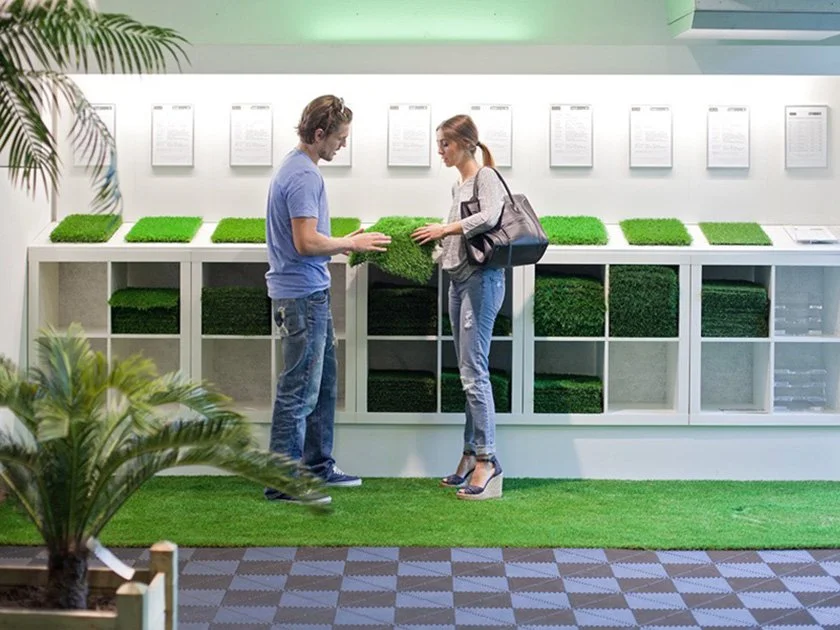 A man and woman are in a store, examining artificial grass samples. The man is holding a small piece of grass while the woman points to her purse. They are standing in front of a display with various green grass samples and informational sheets on th