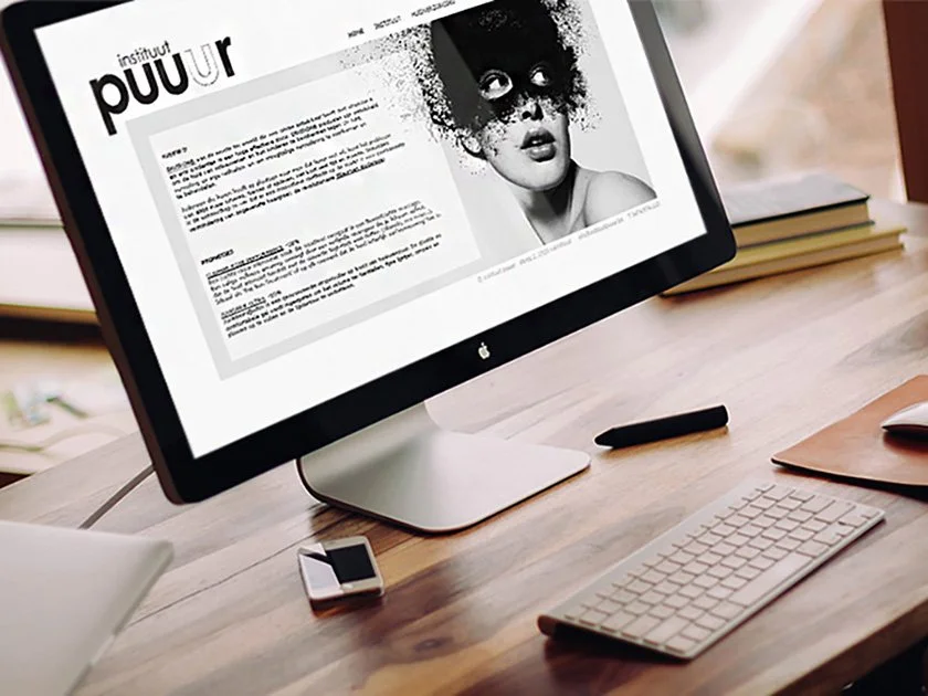 Computer monitor displaying a webpage with the name 'instituut puuur' and an image of a girl with black paint around her eyes, on a wooden desk with a keyboard, smartphone, headphones, and stack of books nearby.