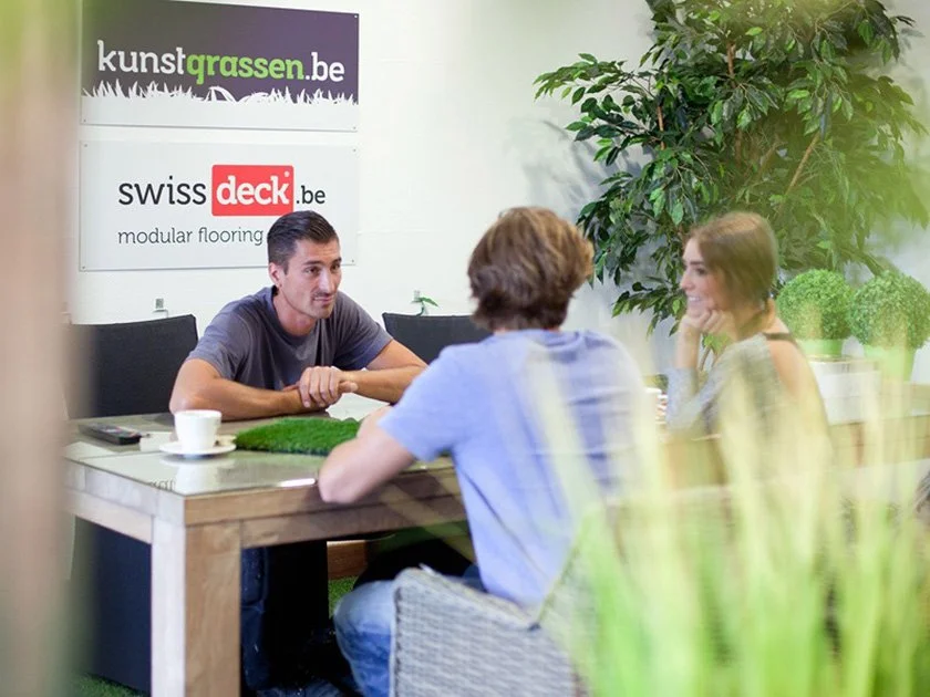 A group of three young adults sitting at a wooden table in an office or meeting room, engaged in conversation. The man on the left is speaking, and two women are listening, one smiling. There are signs on the wall with URLs and text, and a large gree