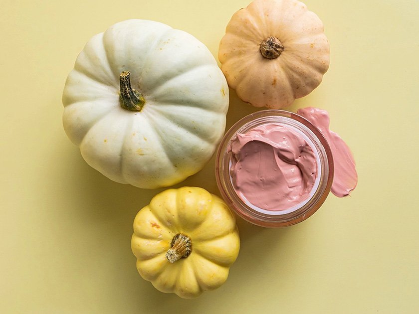 Three decorative mini pumpkins and a jar of pink whipped body butter on a light yellow background.