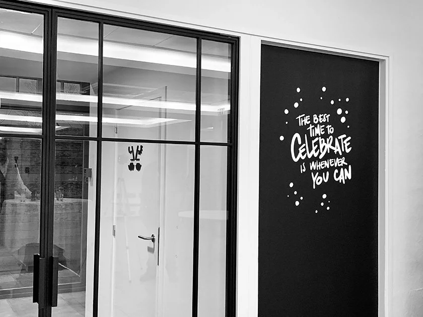 Glass wall with black metal frame, interior office space, and a wall with motivational quote: 'The best time to celebrate is whenever you can', surrounded by dots, in black and white.
