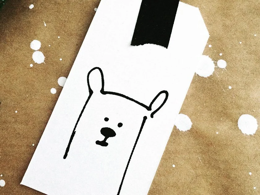 A white tag with a simple black line drawing of a bear's face, attached with a black strip to a brown paper background with white paint splatters.