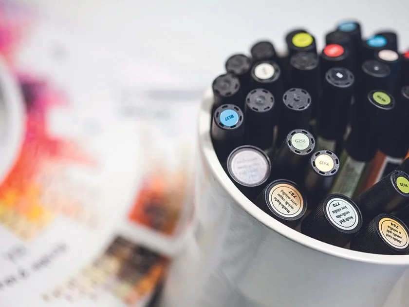Collection of colorful markers in a white cup, with a blurry background of paper.