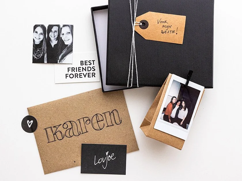 A collection of personalized gift items including a black gift box tied with string, a handwritten tag with "Voor mijn zusje!", black and white photographs of three women, a brown card with the name "Karen" written on it, a black card with "Lojje" wr