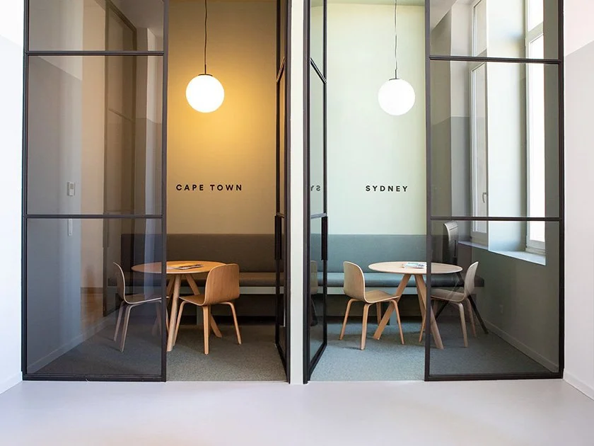Two modern meeting rooms with glass walls, one labeled 'CAPE TOWN' and the other 'SYDNEY,' each with a round table, chairs, and pendant lights.