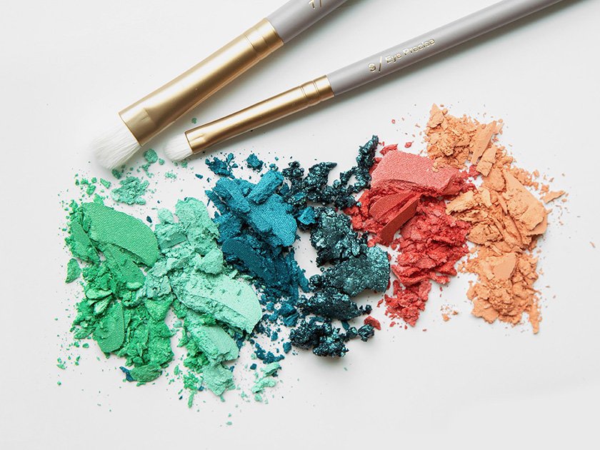 Crushed colorful makeup eyeshadows arranged from green to peach, with two makeup brushes nearby on a white background.