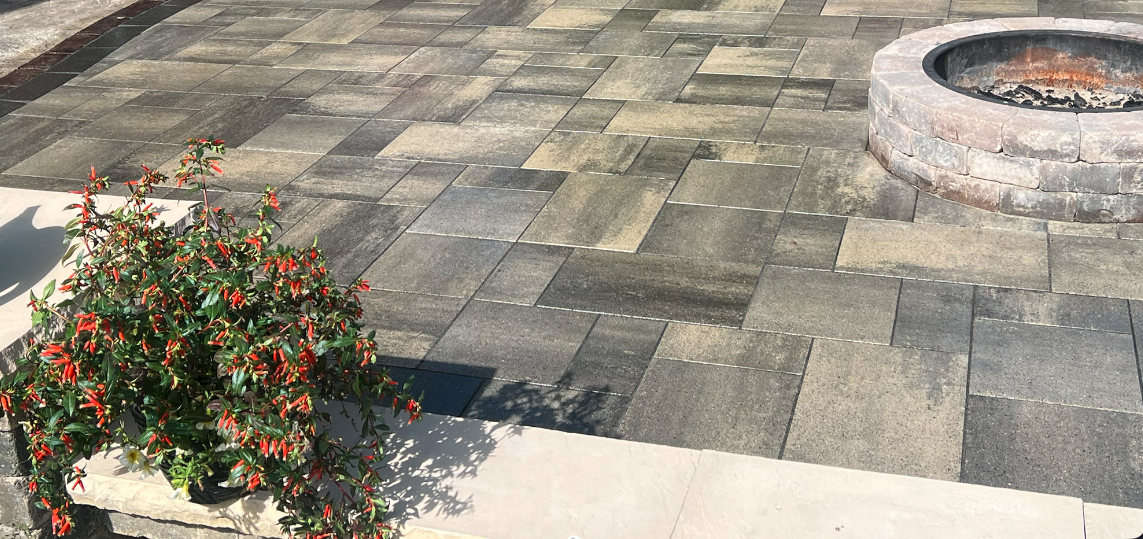 A section of outdoor patio with gray stone pavers, a potted plant with green leaves and red flowers, and a circular brick fire pit.
