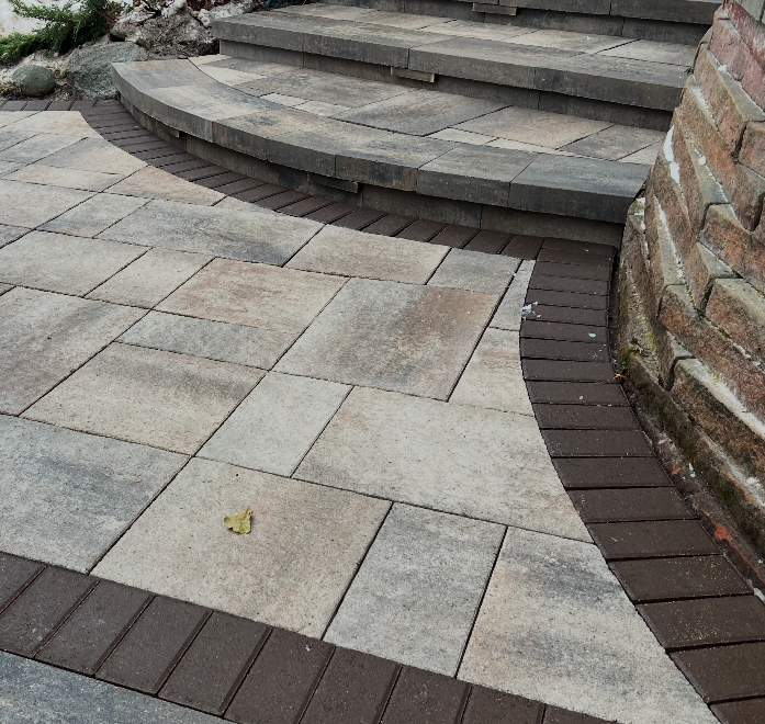 Close-up of a curved stone patio with steps, bordered by a brick wall and a narrow wood strip, outdoors.