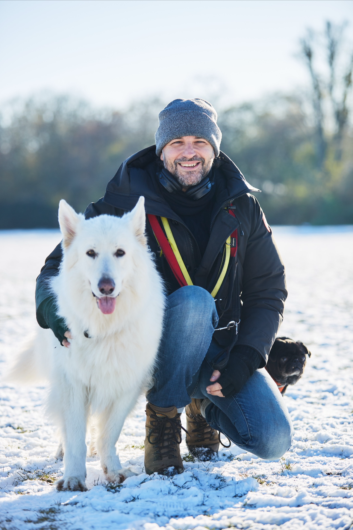 A man kneeling on snow-covered ground with a white dog and a small black dog, winter outdoor scene with trees in the background