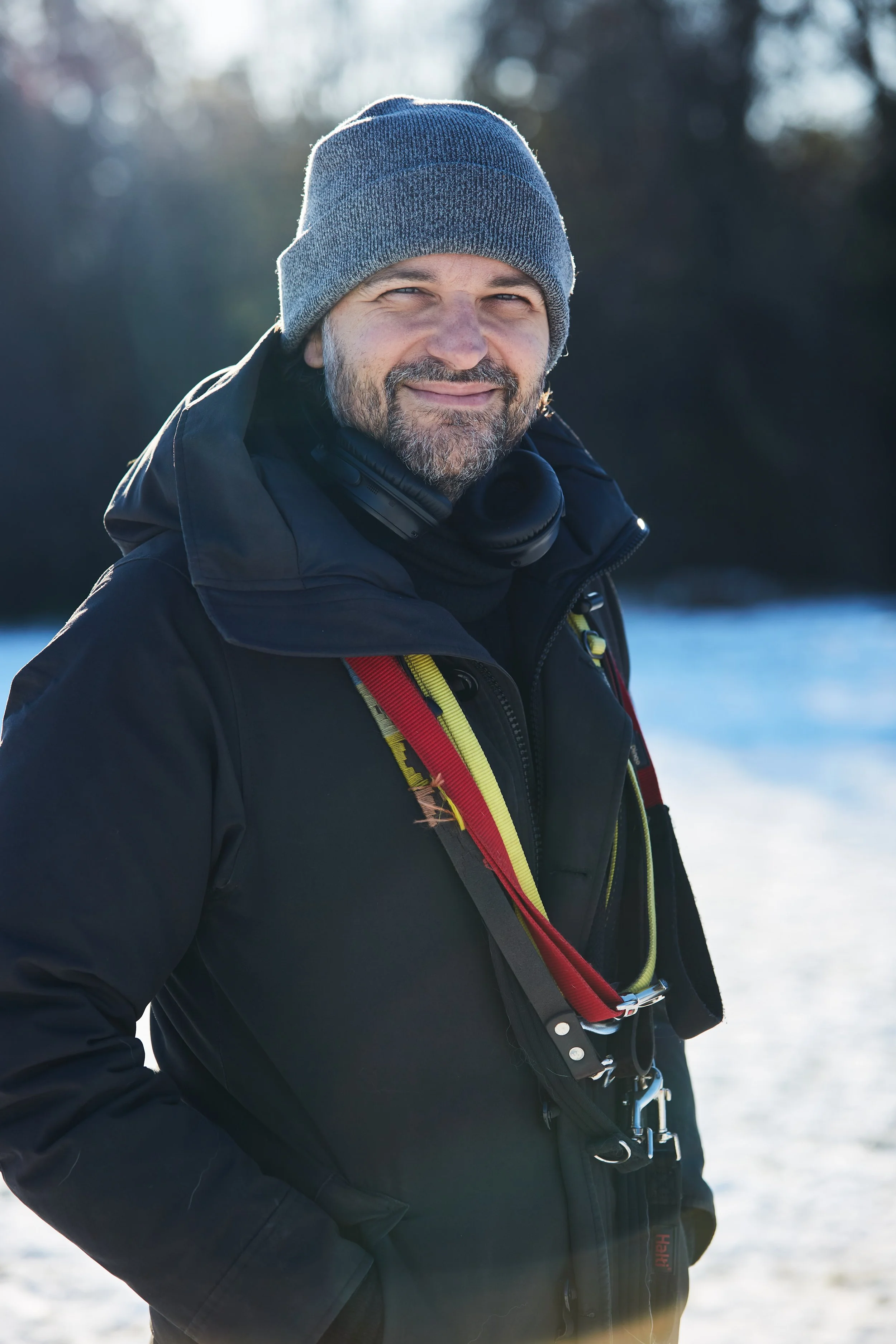 A man dressed in winter clothing standing outdoors in a snowy landscape, wearing a gray beanie, black jacket, and a harness with red and yellow straps, with headphones around his neck, smiling at the camera.