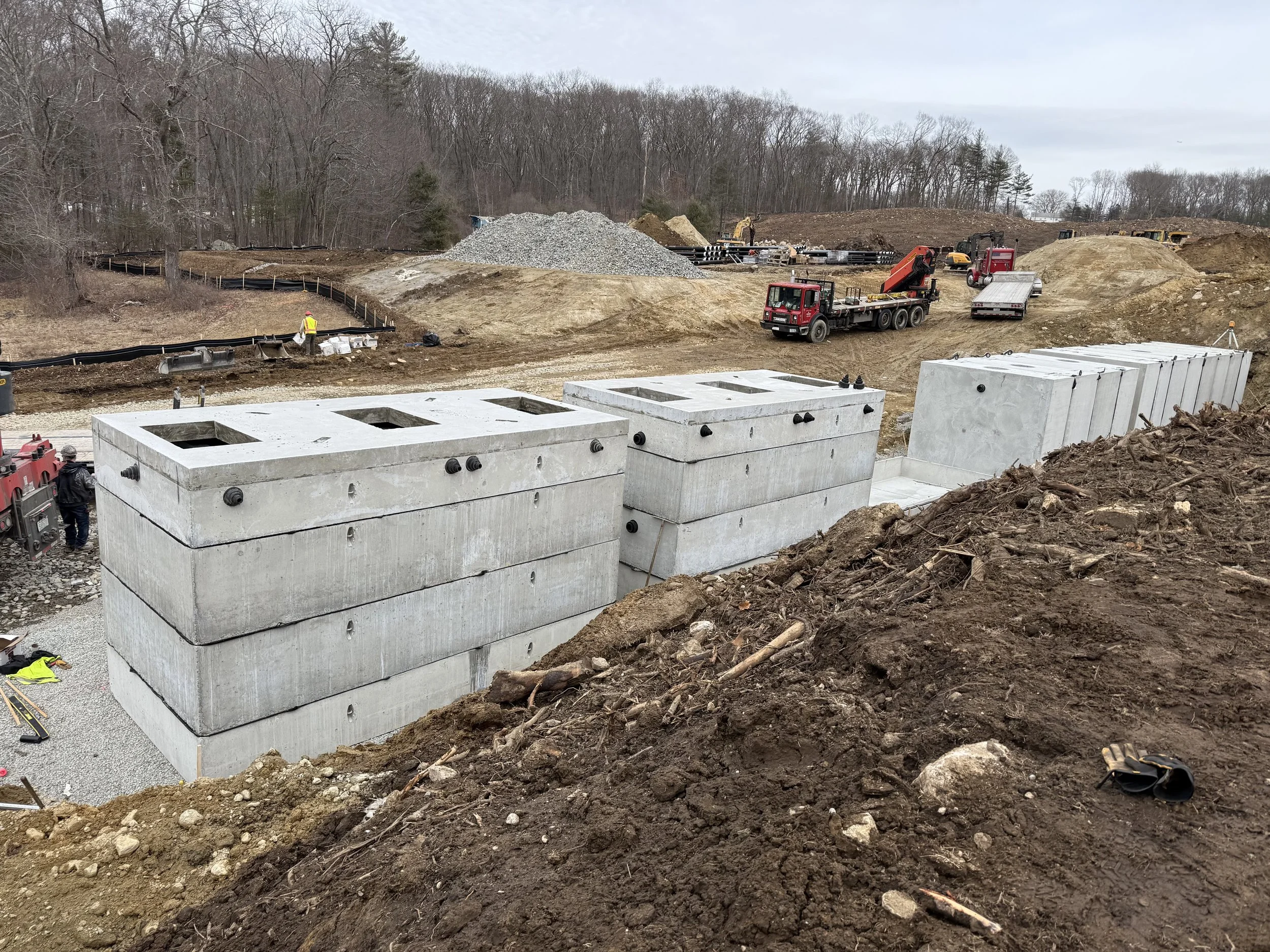 Construction site with large concrete blocks, earth-moving equipment, gravel, and workers preparing the foundation for a building in a wooded area with bare trees. Stormwater and leaching chambers.