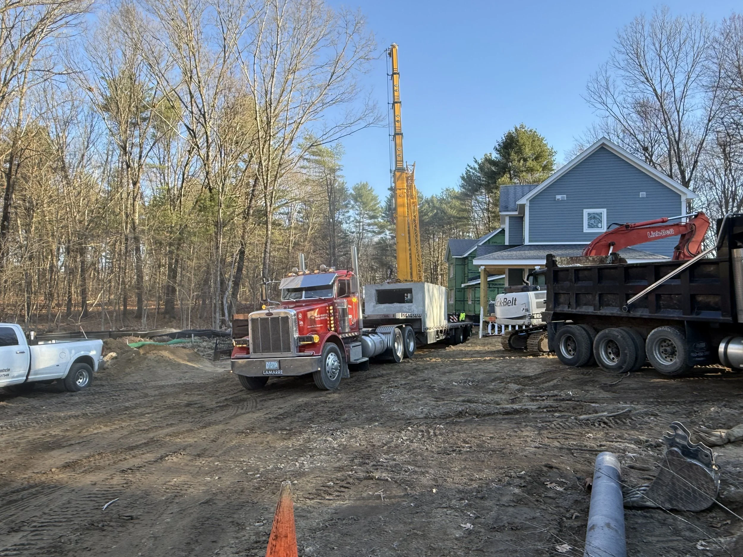 Construction site featuring a large yellow crane lifting materials, with a blue house under construction, surrounded by leafless trees under a clear blue sky. Peterbilt truck carrying precast concrete products.