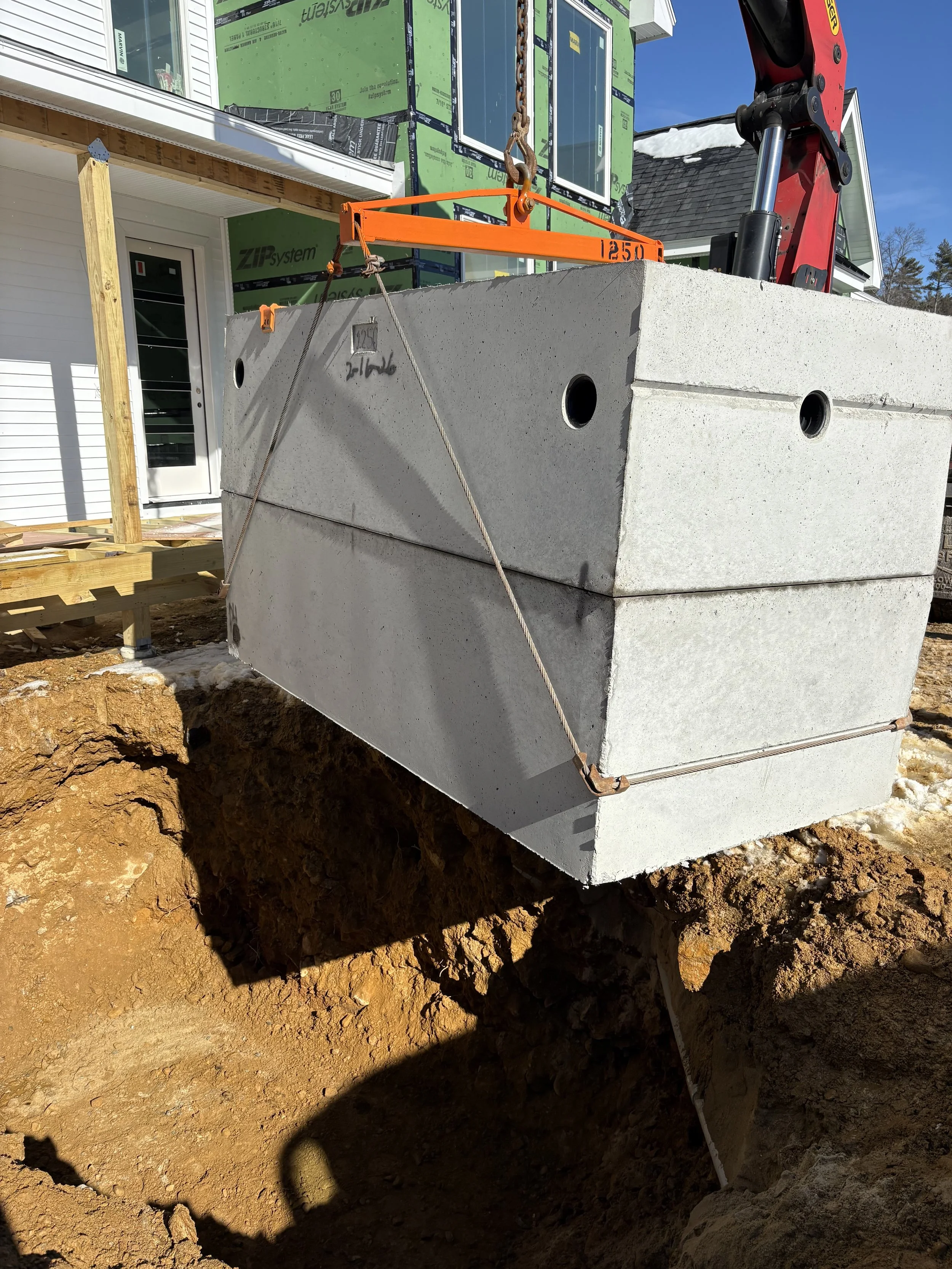 Large concrete block being lowered into a construction site using lifting equipment. The background shows part of a house under construction with green sheathing and a white door. A septic tank is installed.
