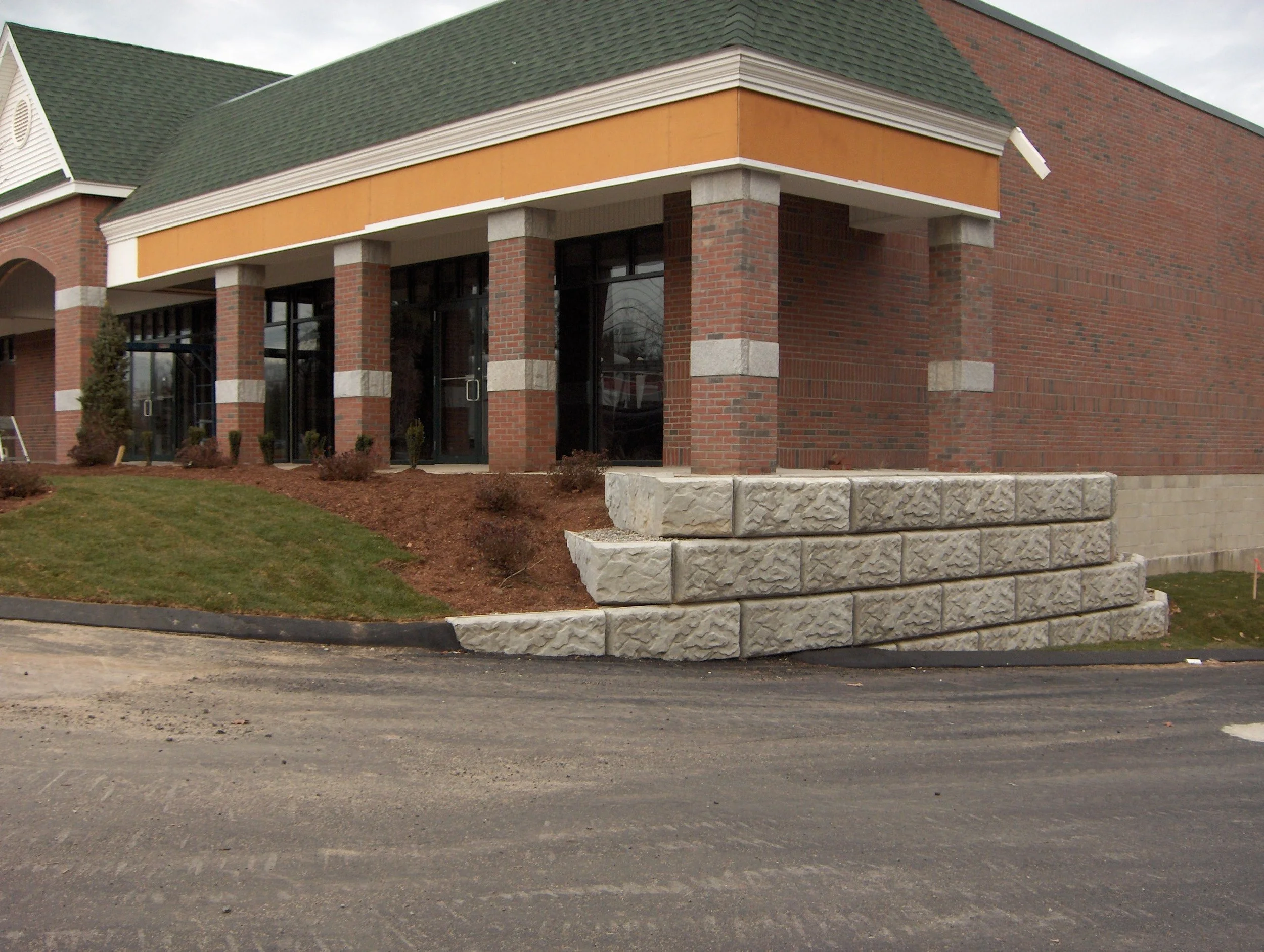 Exterior of a building with brick walls, large windows, a green shingled roof, and a decorative yellow-orange band around the top. There are bushes and a stone curb in front, with a paved parking lot underneath. Wall block installation is completed.