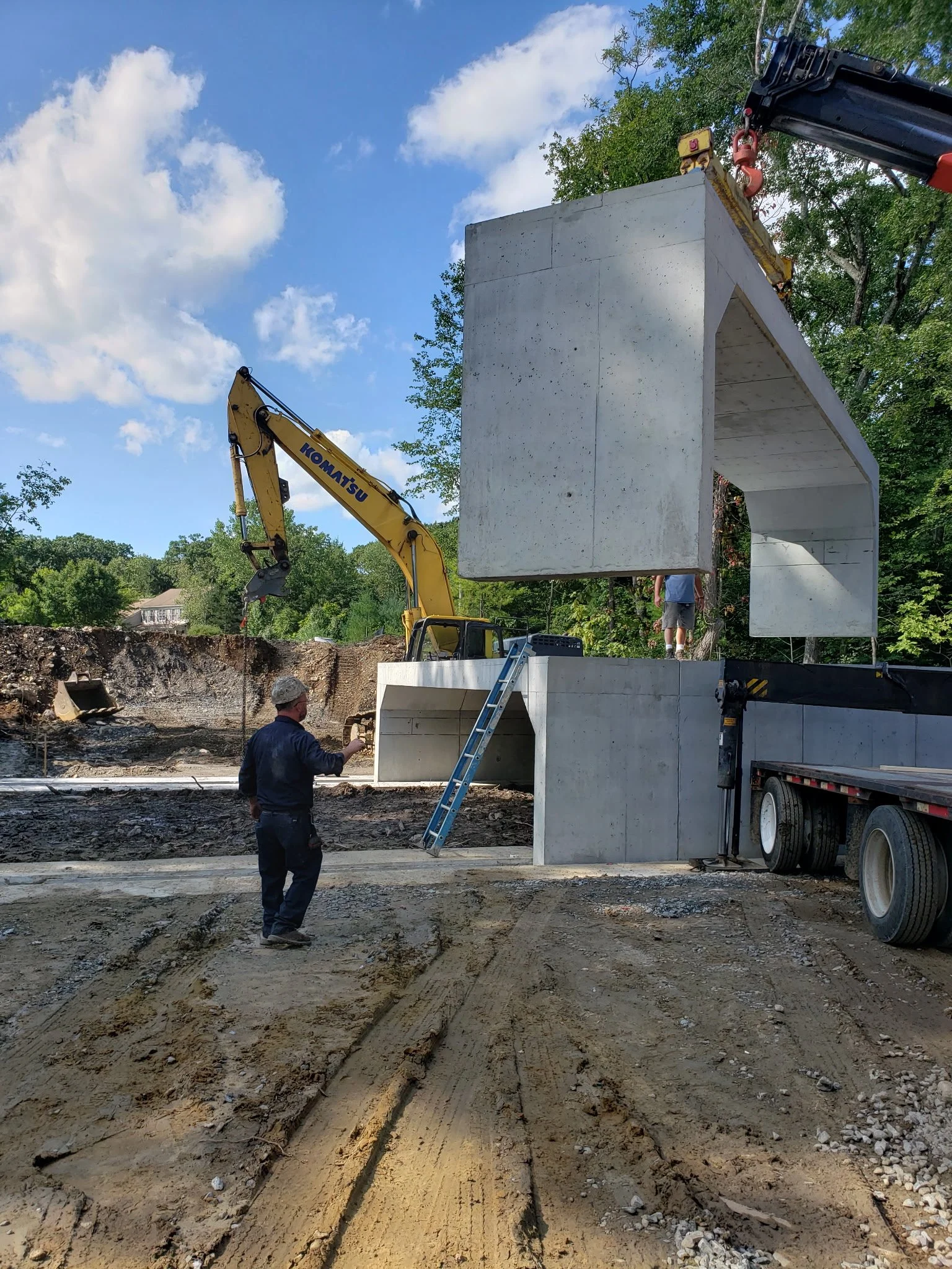 Construction site with large concrete bridge section being set up by crane, workers monitoring, soil and track marks on ground, green trees and blue sky with clouds in background. Precast concrete culvert installation.