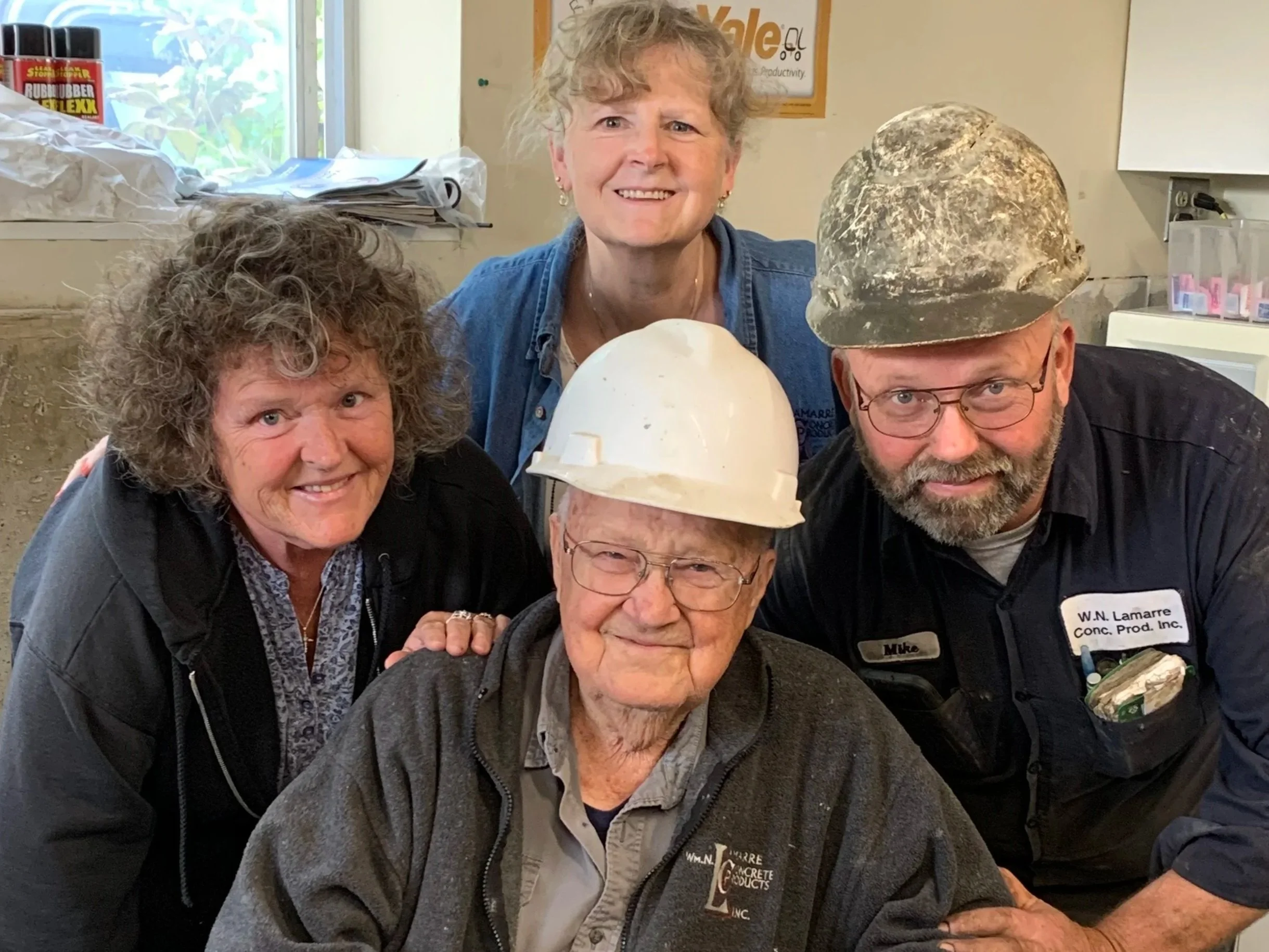 Group of four smiling people, two women and two men, gathered around a table. The older man at the front wearing glasses and a hard hat. The woman on the left has curly hair, and the woman on the back has short blonde hair. The man on the right wears glasses and a dark shirt. They are inside a room with a window, books, and posters visible in the background.
