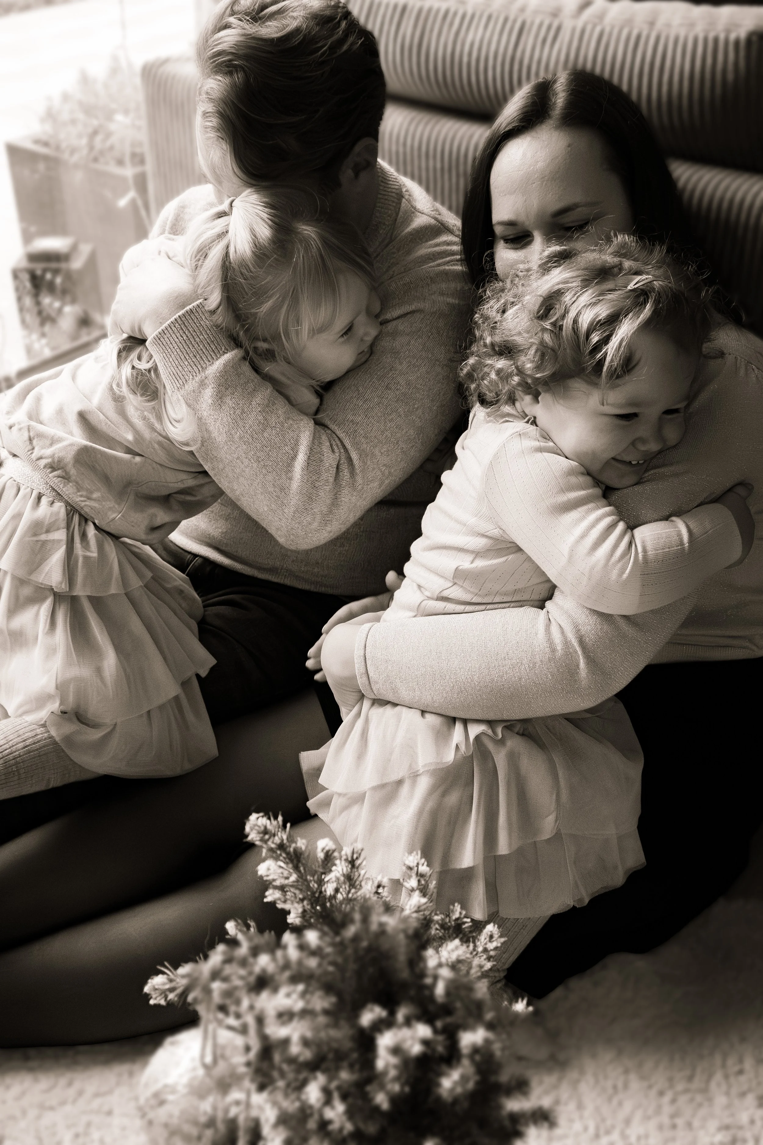 Two women and two young girls hugging and smiling happily indoors near a window with a Christmas tree in the foreground.