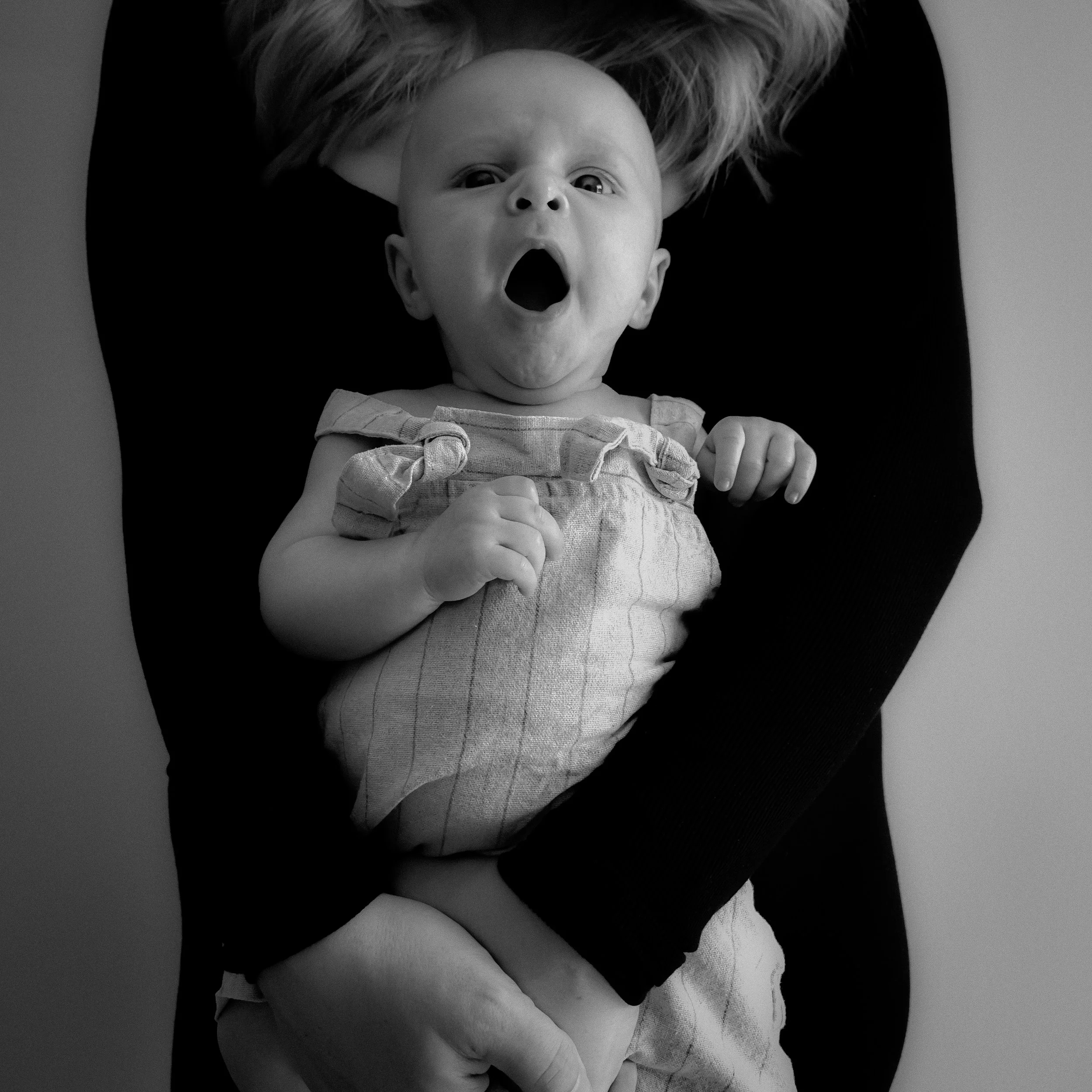 Black and white photo of a baby being held by an adult. The baby is lying on their back with their mouth open, eyes wide, and looking up. The adult's arms are visible, holding the baby securely.