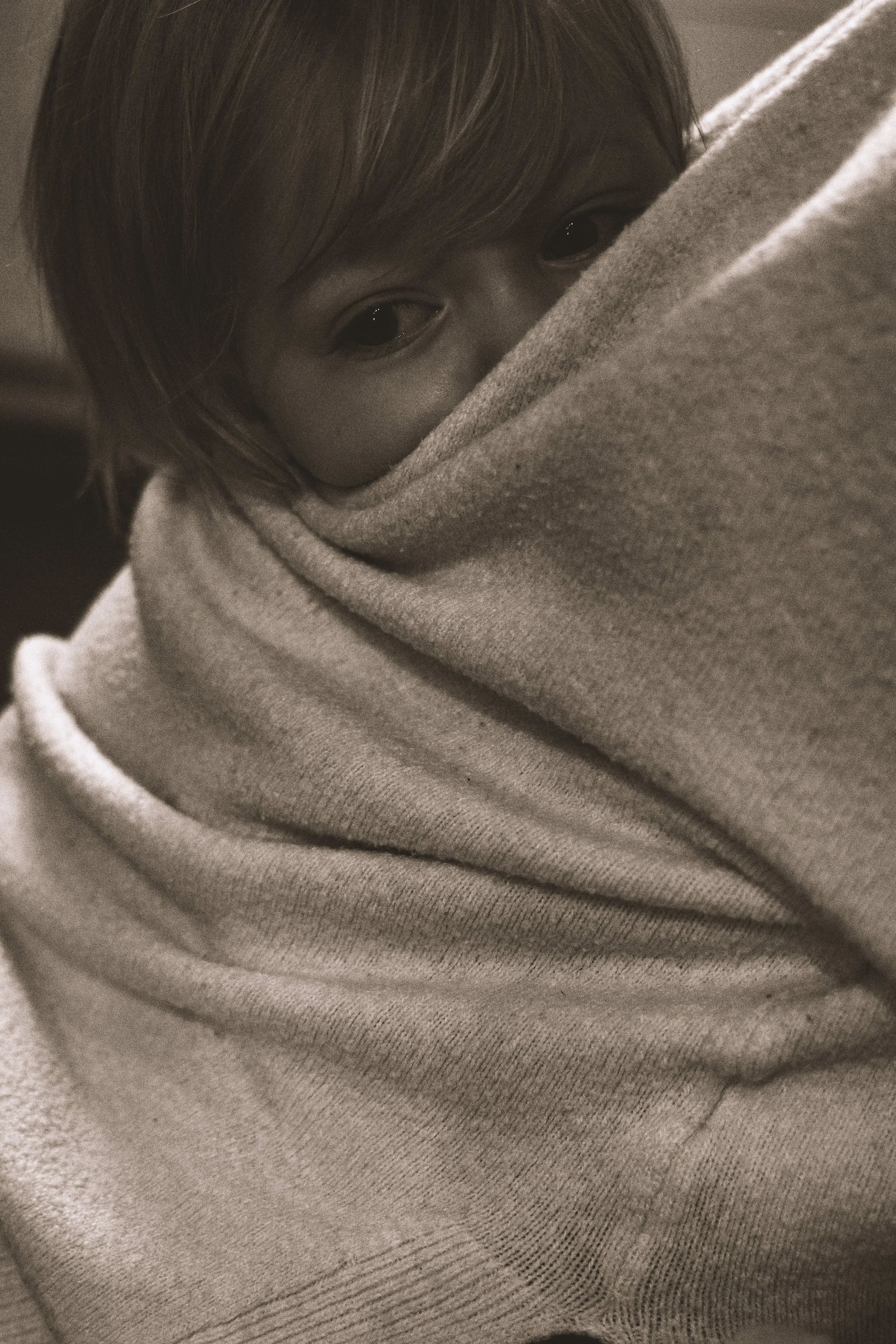 A person with short hair wraps a blanket or cloth around their face, revealing only their eyes looking directly at the camera.