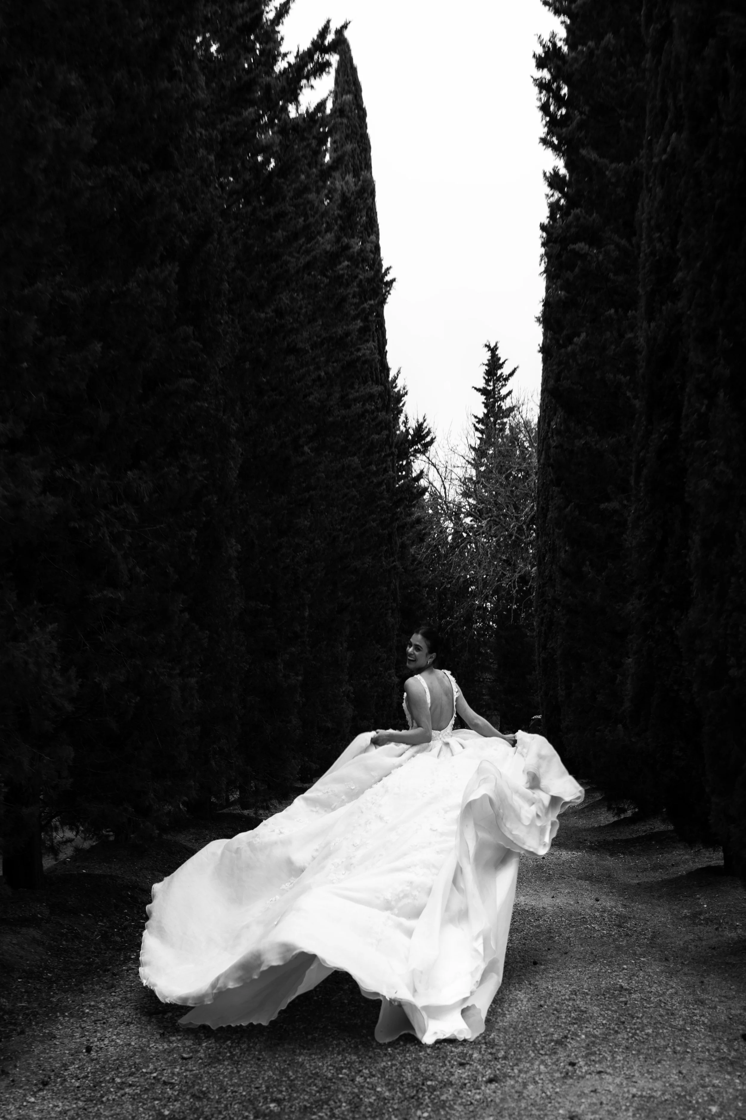 Braut in weißem Hochzeitskleid, die im Freien zwischen hohen, dunklen Bäumen dreht, lächelnd und in Bewegung, im Schwarz-Weiß-Foto