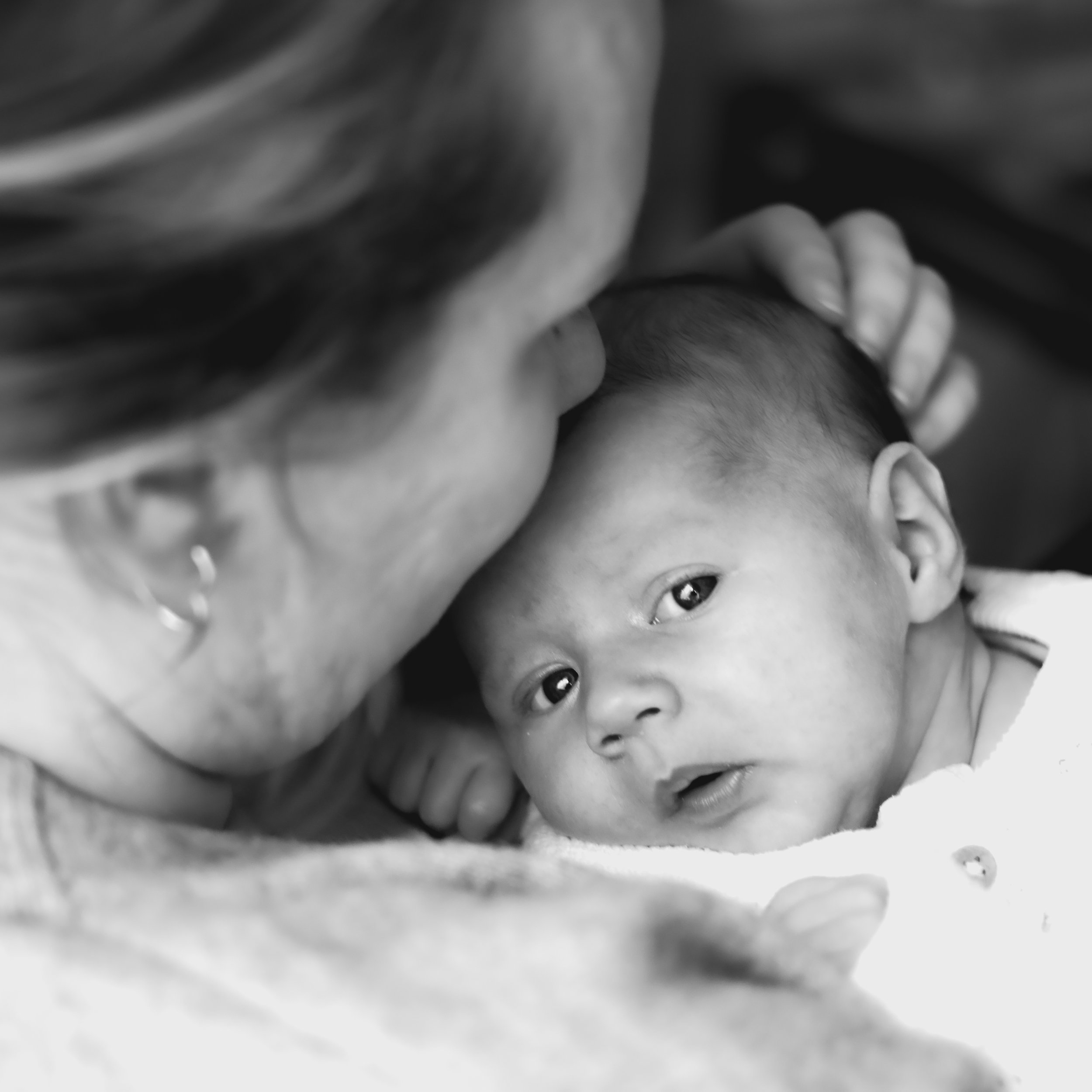 A woman holding a baby close, with her face near the baby's head, in a tender moment. The baby is looking up with wide eyes.