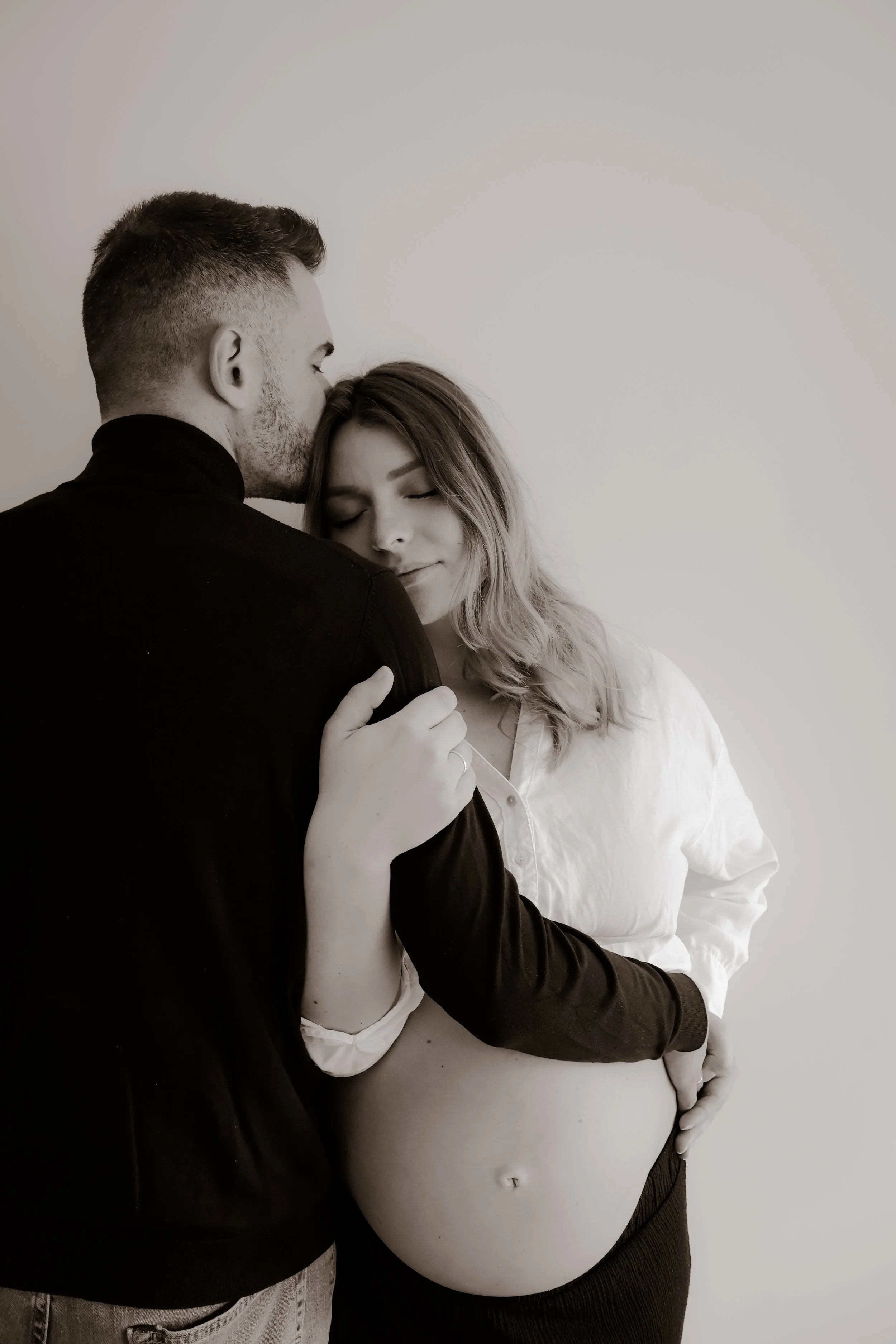 A man and pregnant woman embrace gently, with the man kissing her forehead and the woman smiling with her eyes closed, against a plain background.