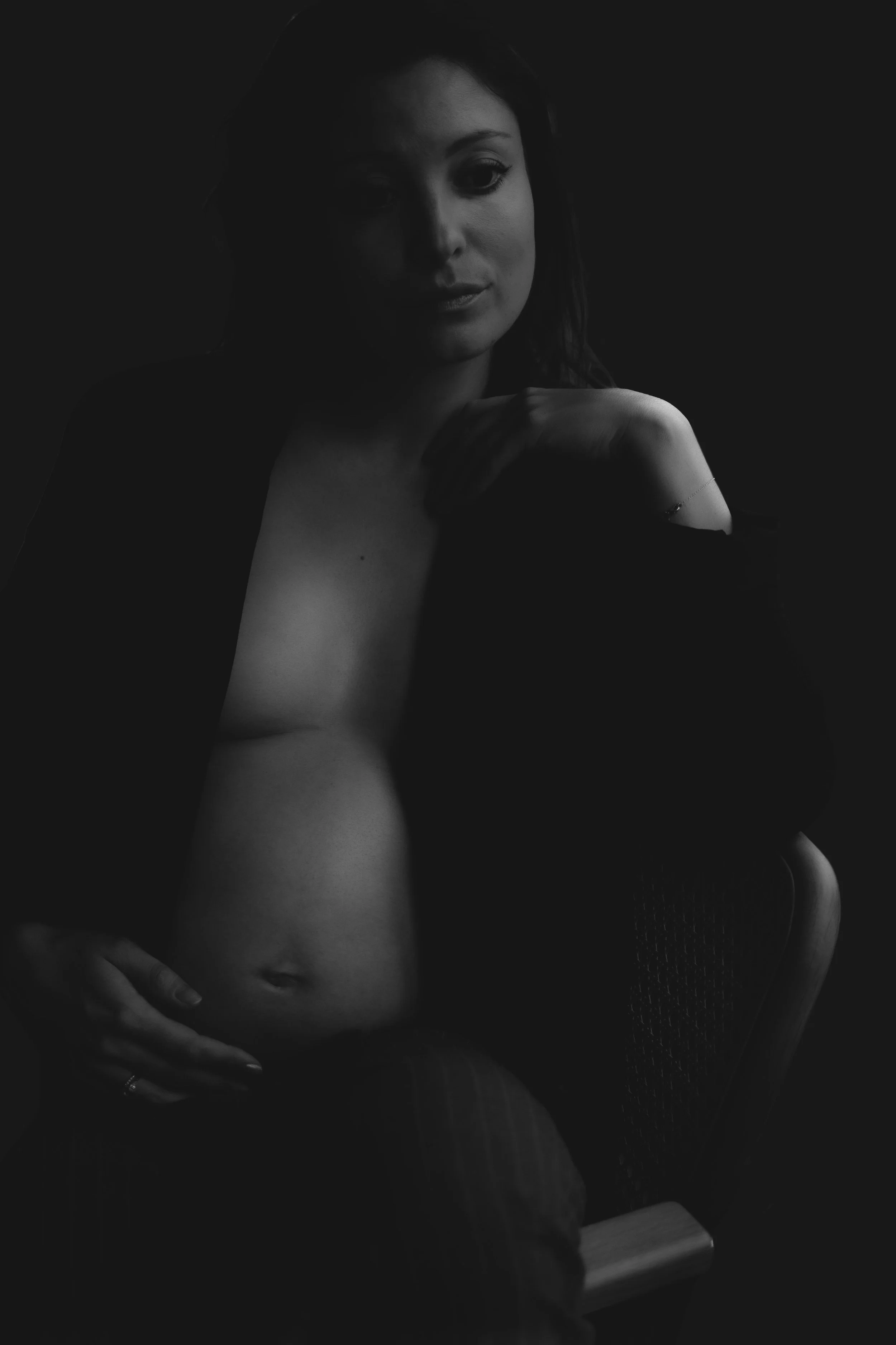 A woman with dark hair sitting in a chair, with her hand resting on her shoulder. The image is in black and white, with shadows partially obscuring her face and body.