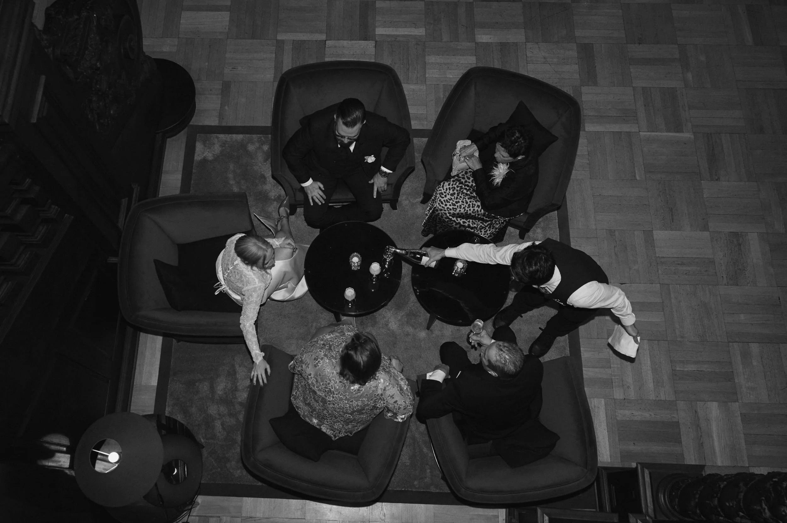 A group of six adults socializing around a small round table in a living room or lounge area, with some seated on couches and others standing. The scene is captured from above in black and white.