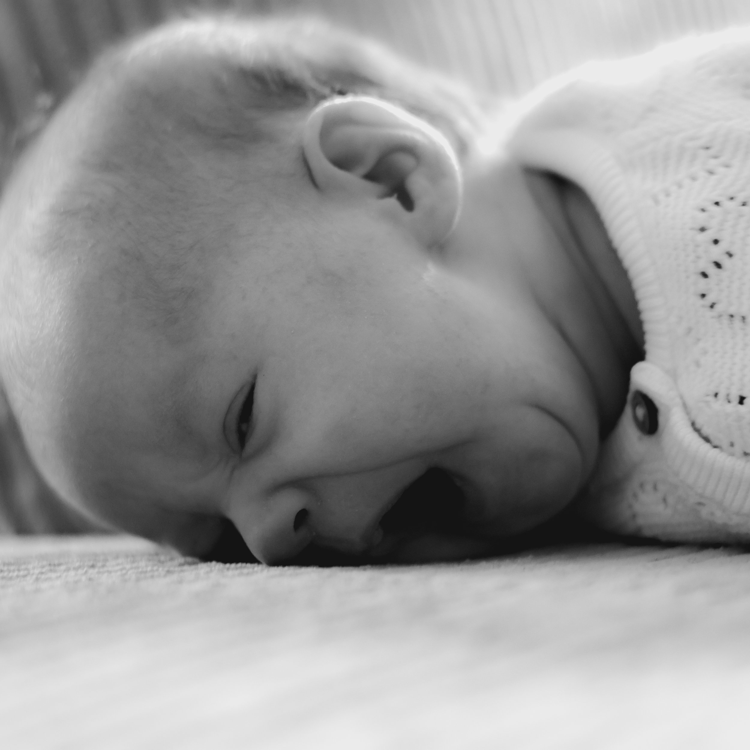 Close-up black-and-white photo of a sleeping baby's face, lying on a surface with eyes partially open, wearing a knitted garment.