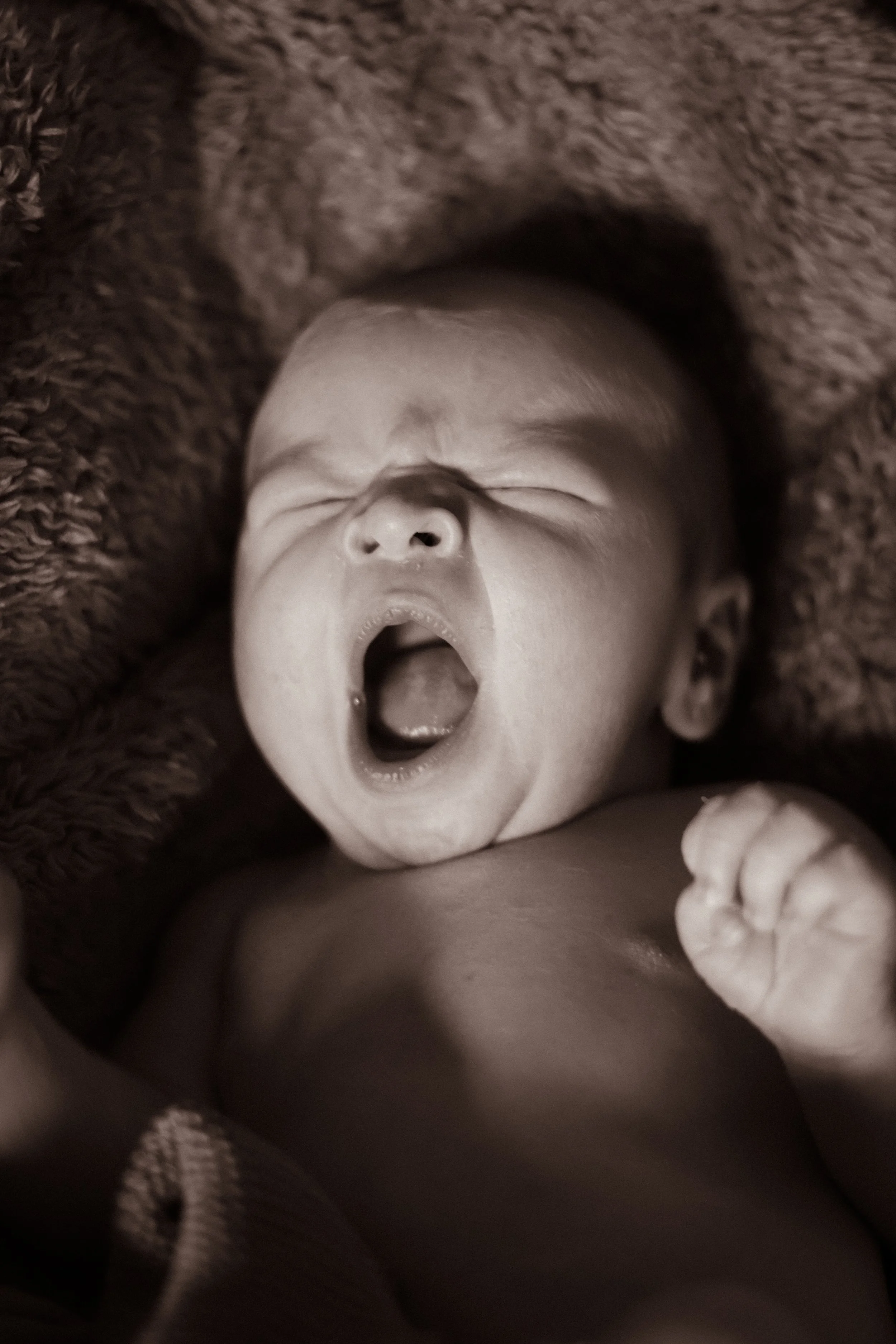 A baby lying on a soft, textured blanket, yawning with eyes closed and mouth wide open.