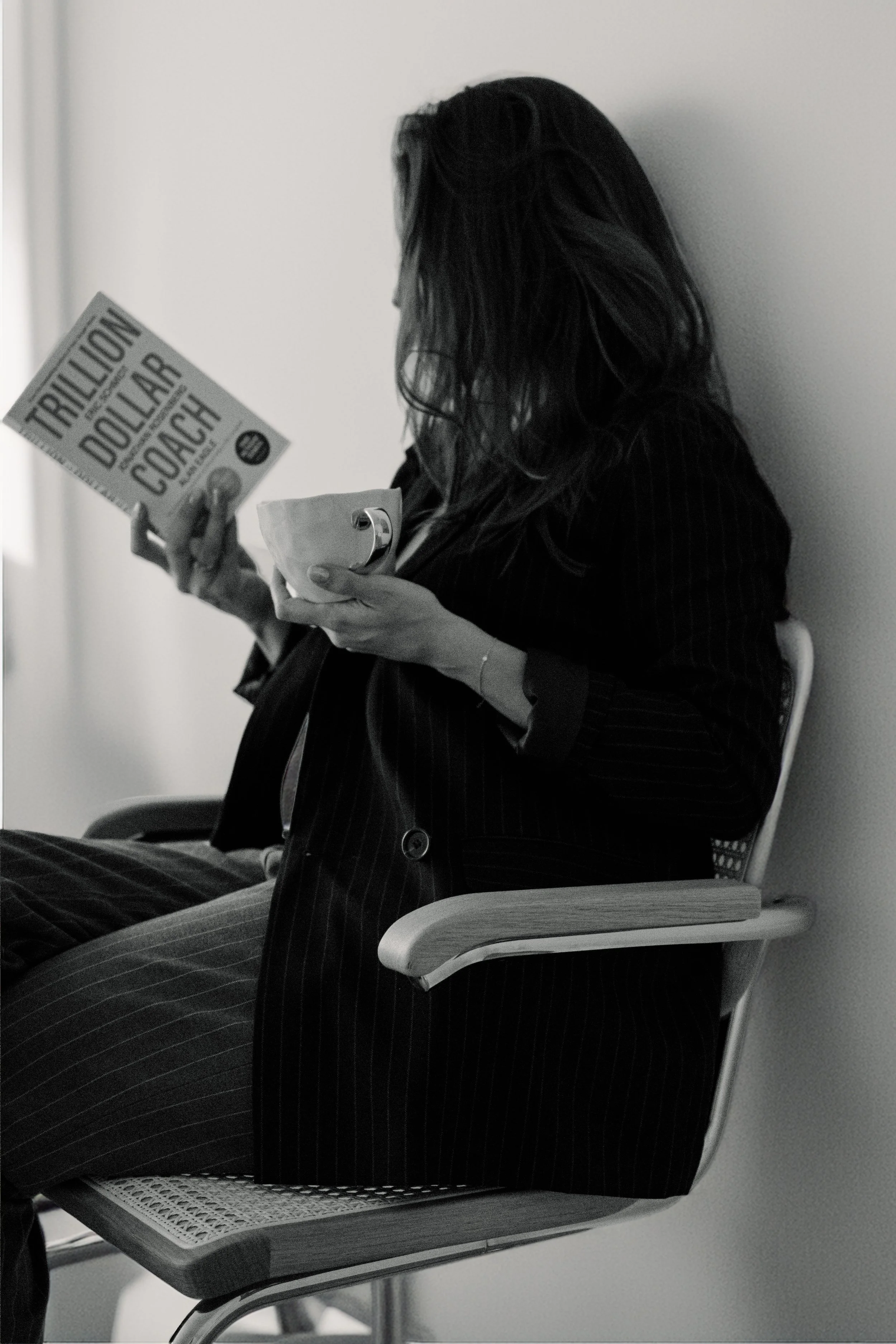A woman with long hair dressed in pinstripe suit sitting on a chair reading a book titled "Trillion Dollar Coach".