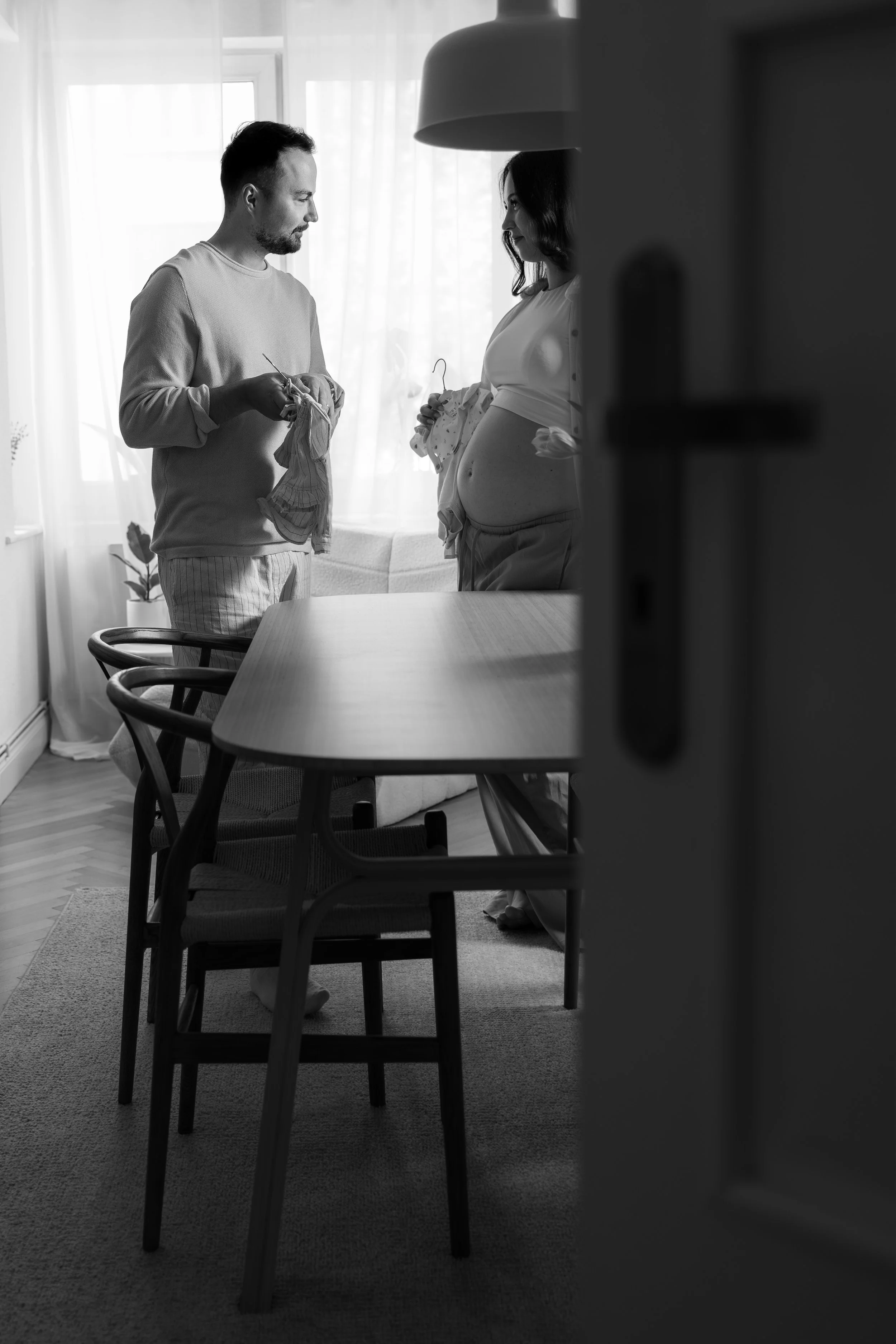 Schwarzweißfoto von einem schwangerschaftlichen Paar im Wohnzimmer, sie hält Babyklamotten, er hält Socken. Sie stehen gegenüber, im Hintergrund große Fenster mit Vorhängen, im Vordergrund Möbel, ein Tisch mit Stühlen.
