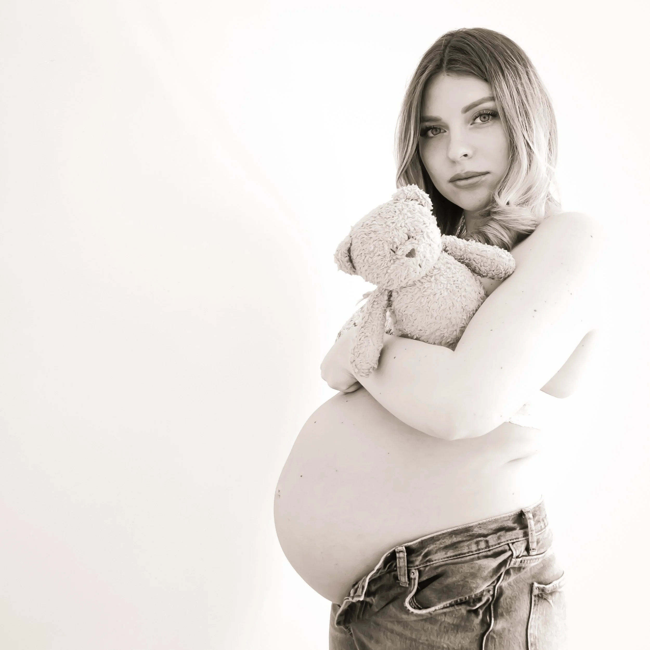 A pregnant woman with shoulder-length hair holding a teddy bear against a plain light background.