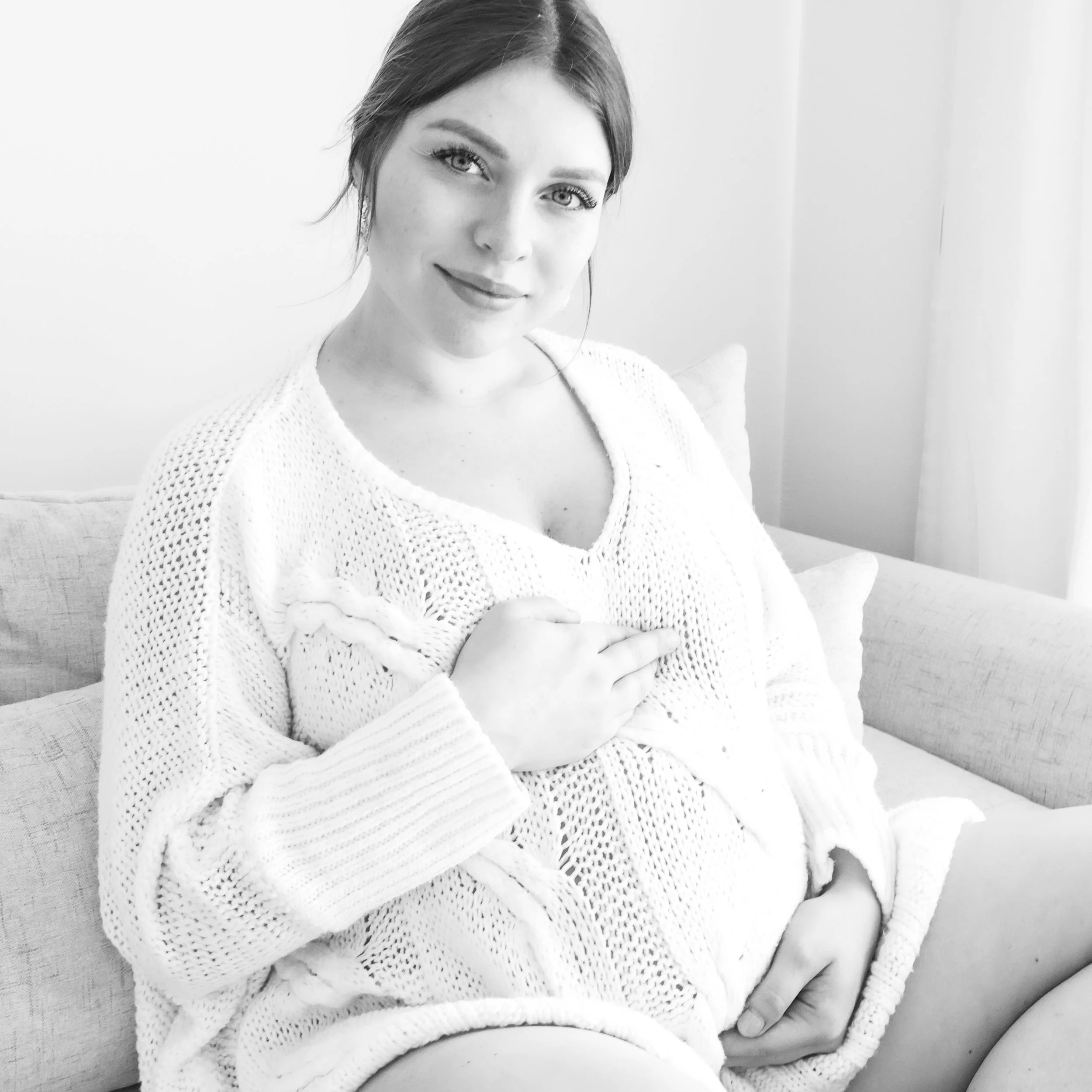 A woman sitting on a couch, smiling, wearing a knit sweater, with her hand over her chest in a cozy, well-lit room.