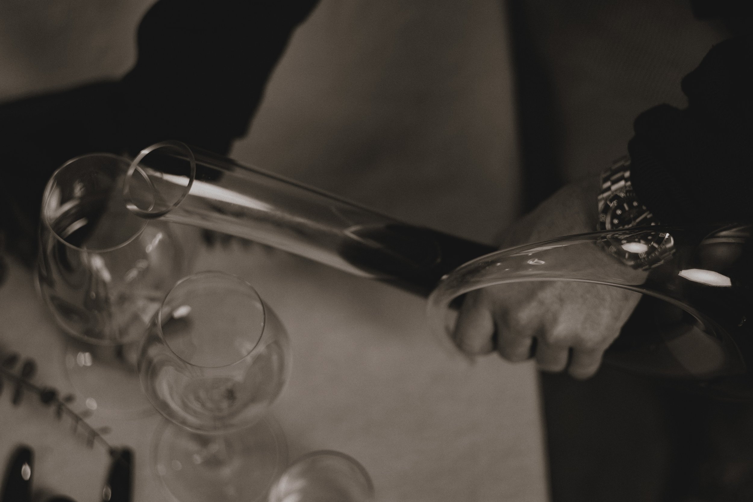 A person wearing black gloves pouring wine from a bottle into multiple empty glasses on a table.