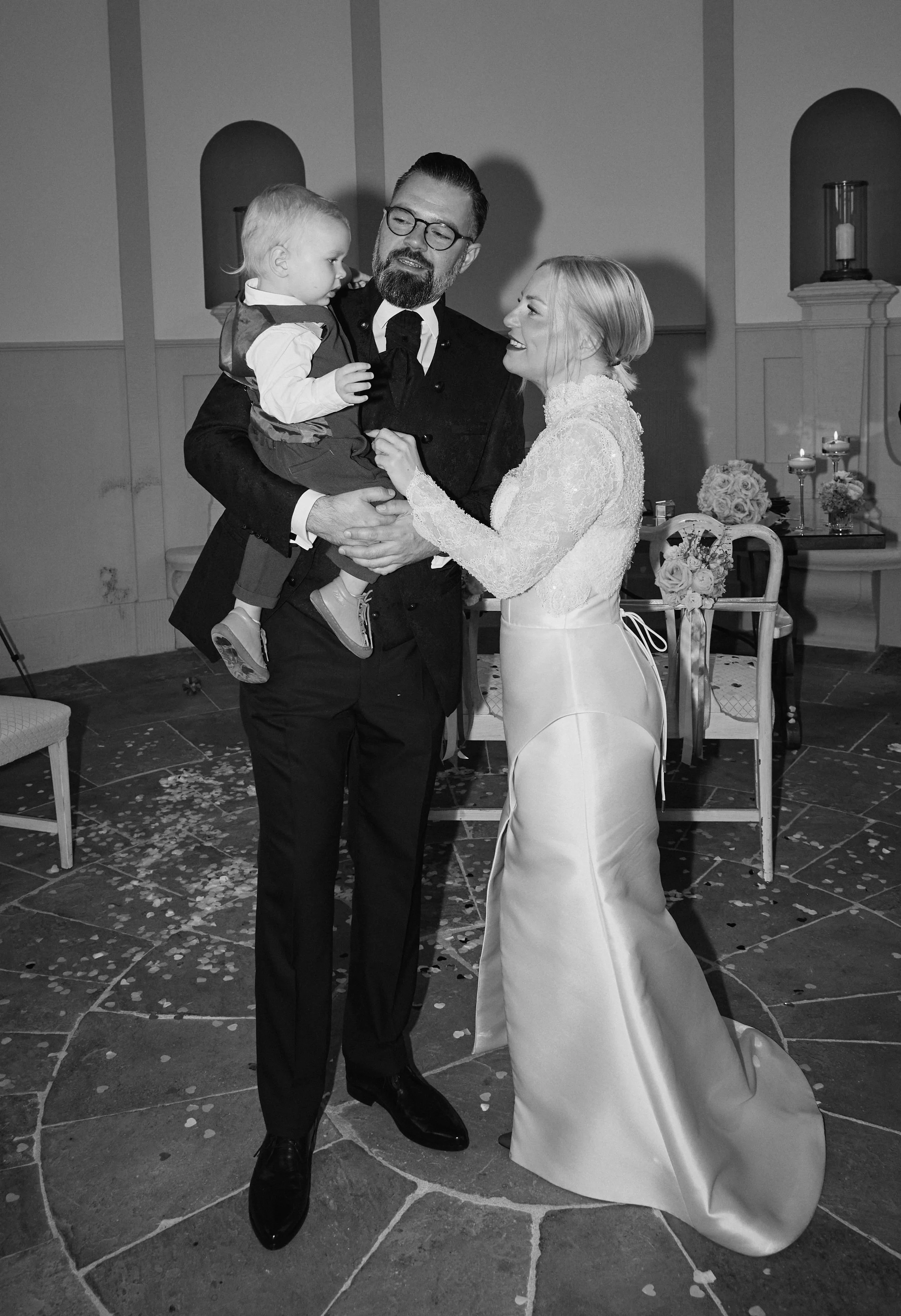 Black and white photo of a wedding scene with a bride, groom, and a young child. The groom holds the child, and the bride stands next to them. The background features candles, flowers, and wedding decorations.