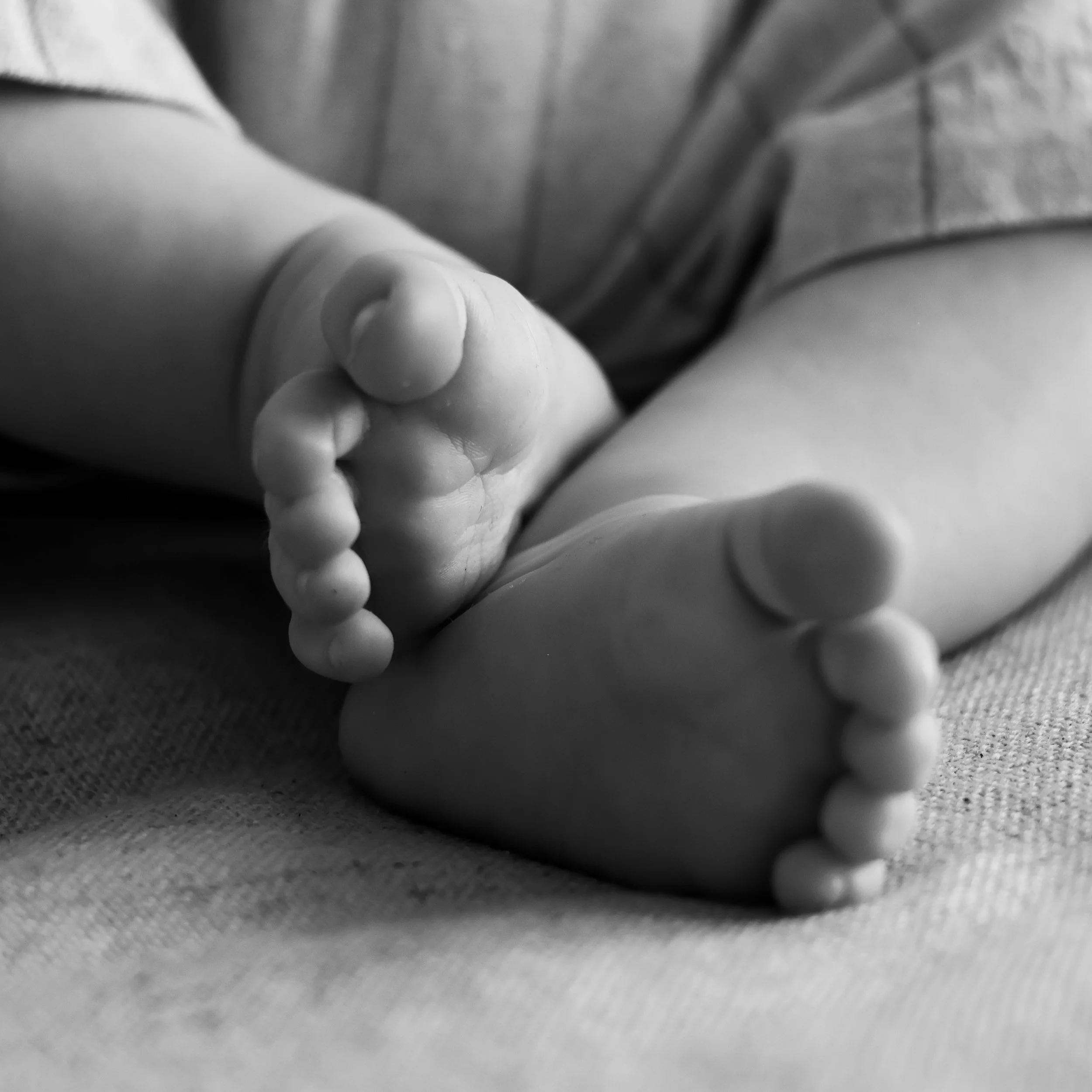 A close-up black-and-white photograph of a baby’s small feet resting on a soft fabric surface, with the focus on the toes.
