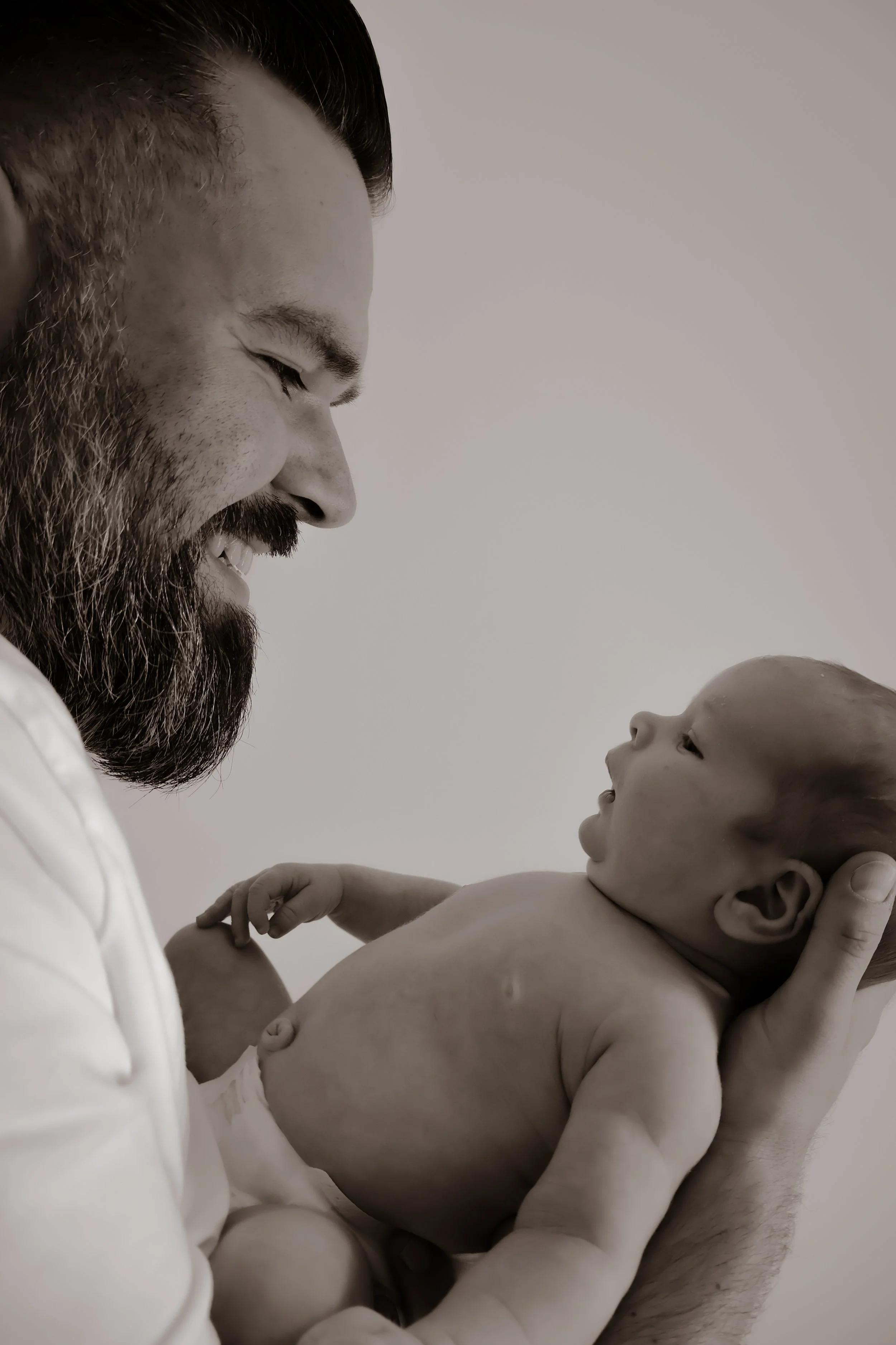A bearded man holding a baby in his arms, both smiling and looking at each other.