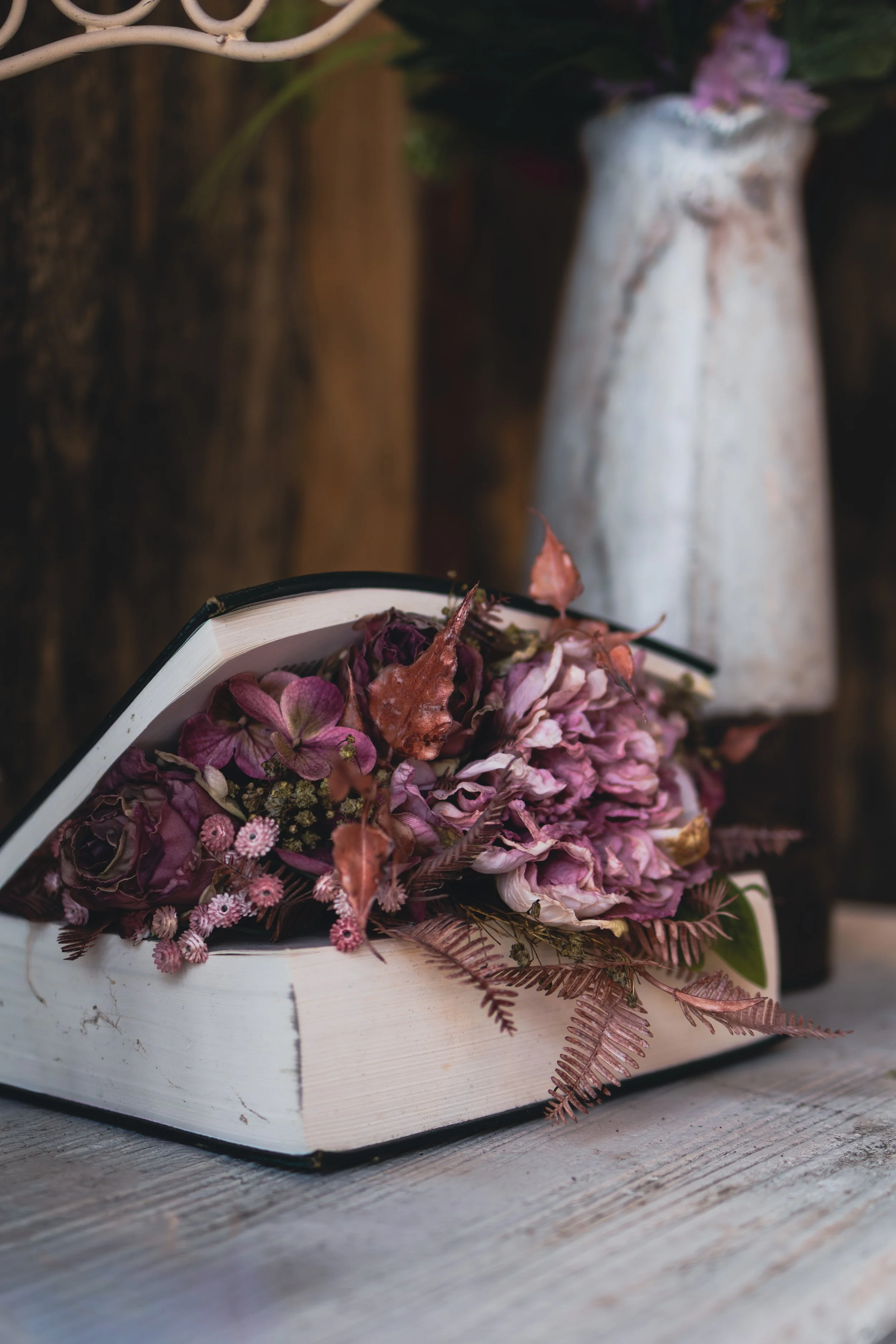 Ein offenes Buch, das mit getrockneten rosa und lilafarbenen Blumen und Blättern dekoriert ist, steht auf einem Holztisch, im Hintergrund eine weiße Vase mit Blättern und einer Blume.
