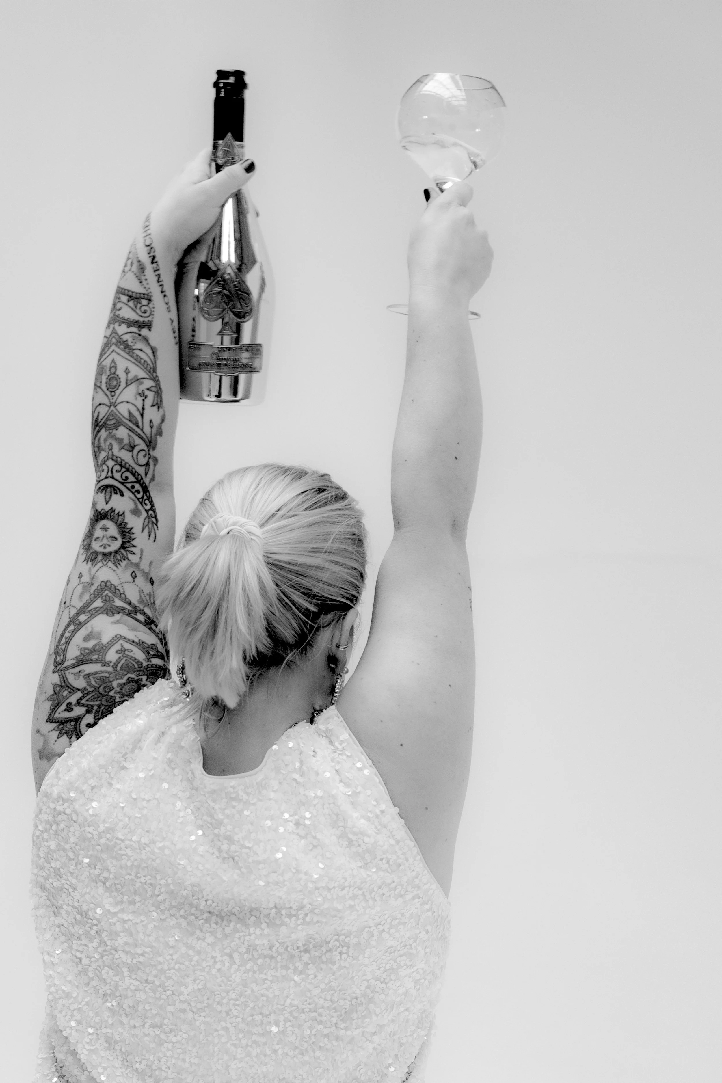 A woman with blond hair tied back, wearing a sequined dress, holding a bottle of champagne in one hand and a wine glass in the other, raising them in celebration.