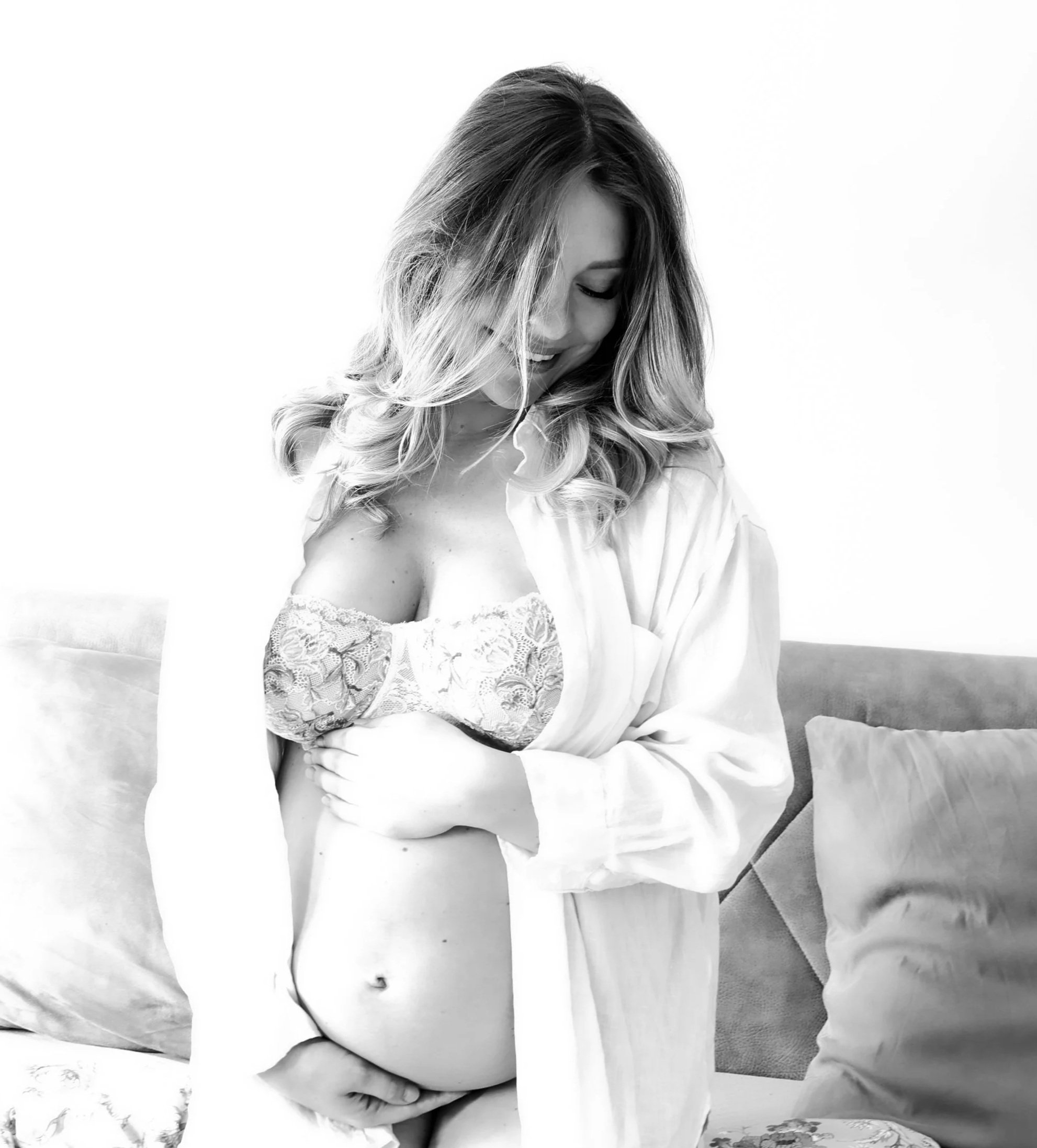 A woman with long wavy hair smiling and looking down, wearing a lace bra and a loose shirt that falls off her shoulder, sitting on a sofa with pillows, in black and white.