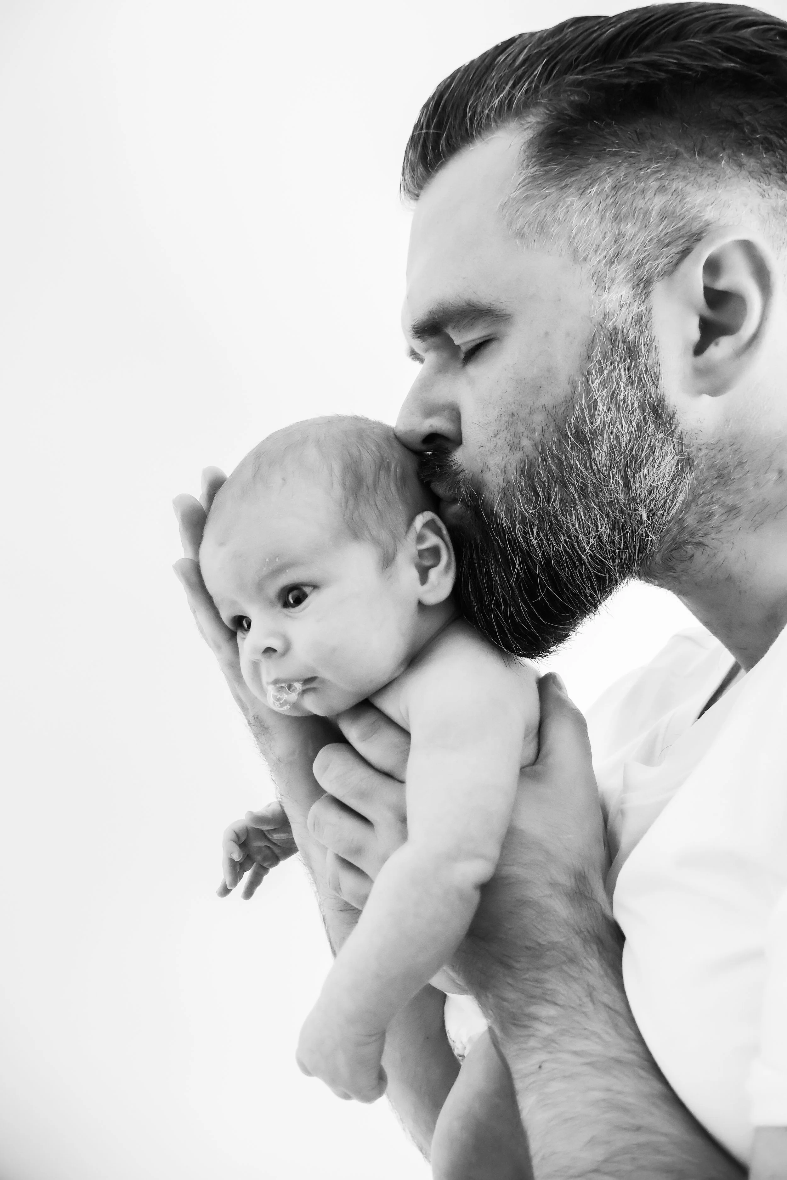 A man with a beard gently kisses an infant on the forehead, holding the baby close in a tender moment.