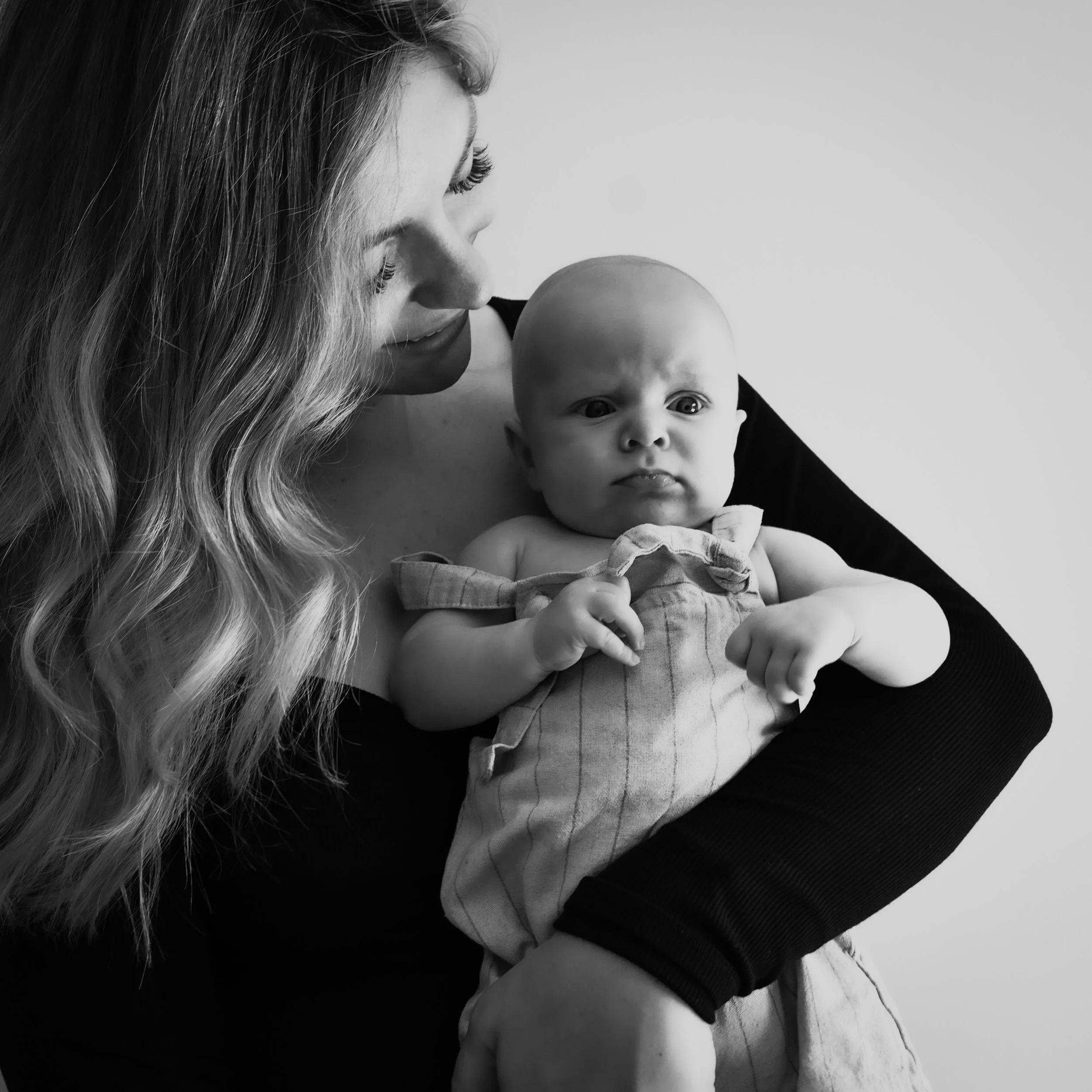 A woman holding a baby, both looking at each other. The woman has wavy hair and is wearing a black top. The baby appears angry or confused, holding a small object, wearing a striped outfit.