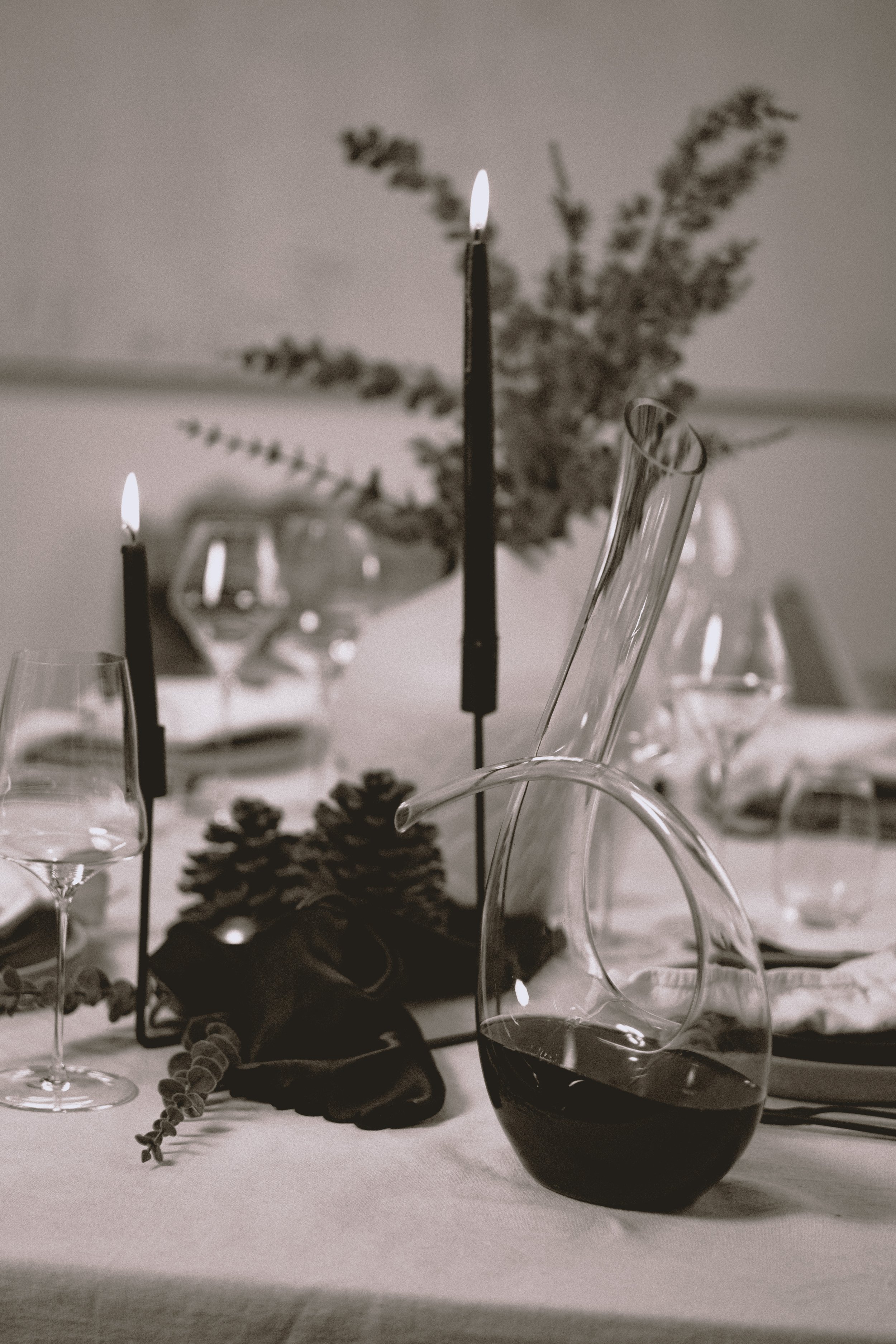 Ein gedeckter Tisch mit Weingläsern, einem Weinballon, Kerzen, Tannenzapfen, Dekorationen und einer Flasche Rotwein in Schwarz-Weiß. Im Hintergrund sind getrocknete Pflanzen zu sehen.