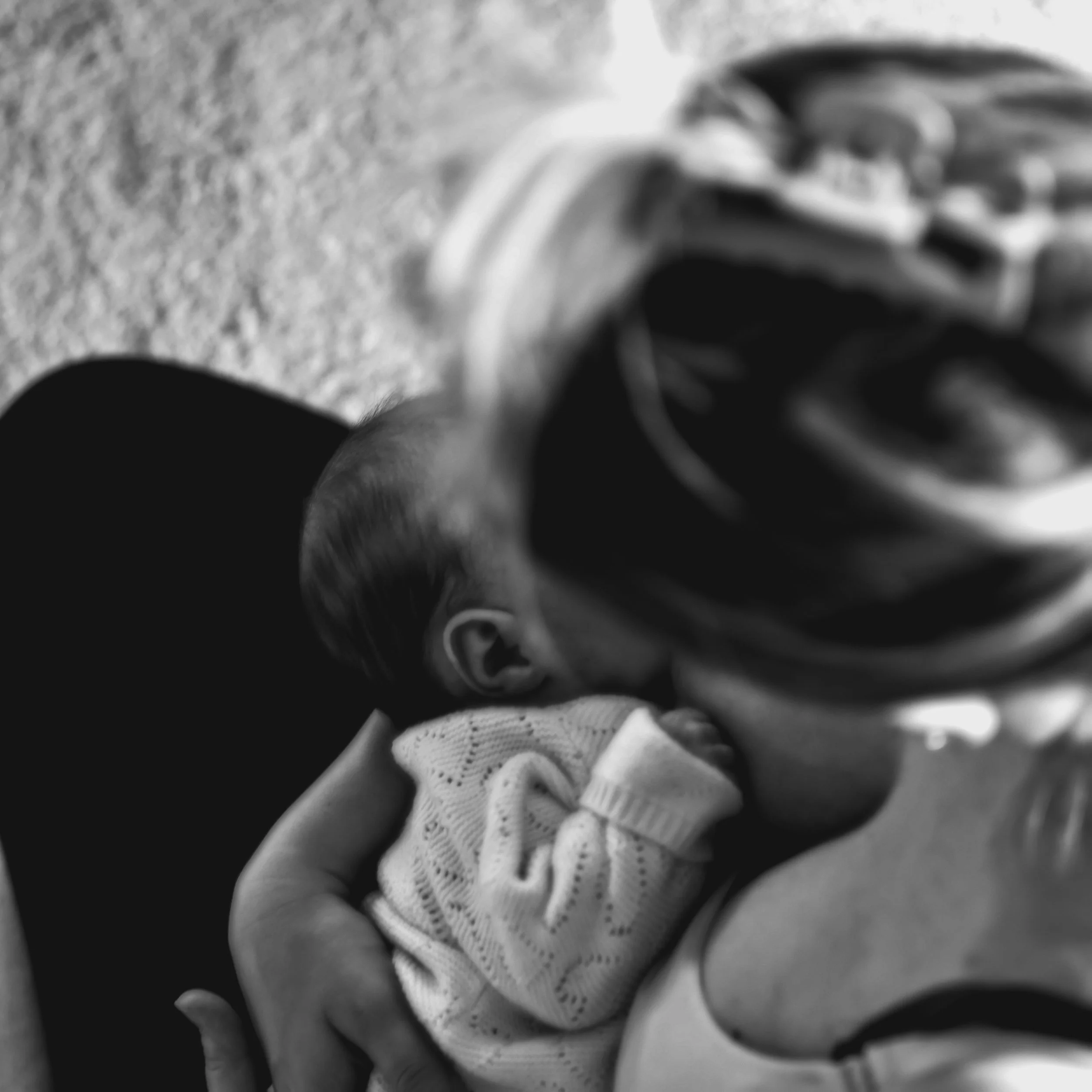 A close-up black and white photo of a person holding a sleeping baby wrapped in a blanket, with the baby's face resting on their chest.