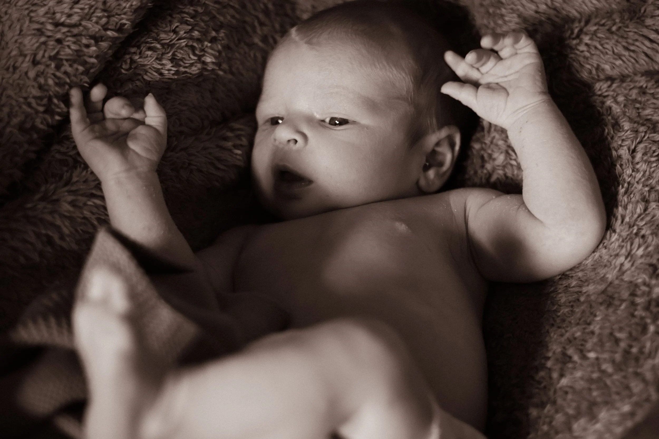 A newborn baby lying on a soft blanket, with arms raised and mouth slightly open, captured in black and white.