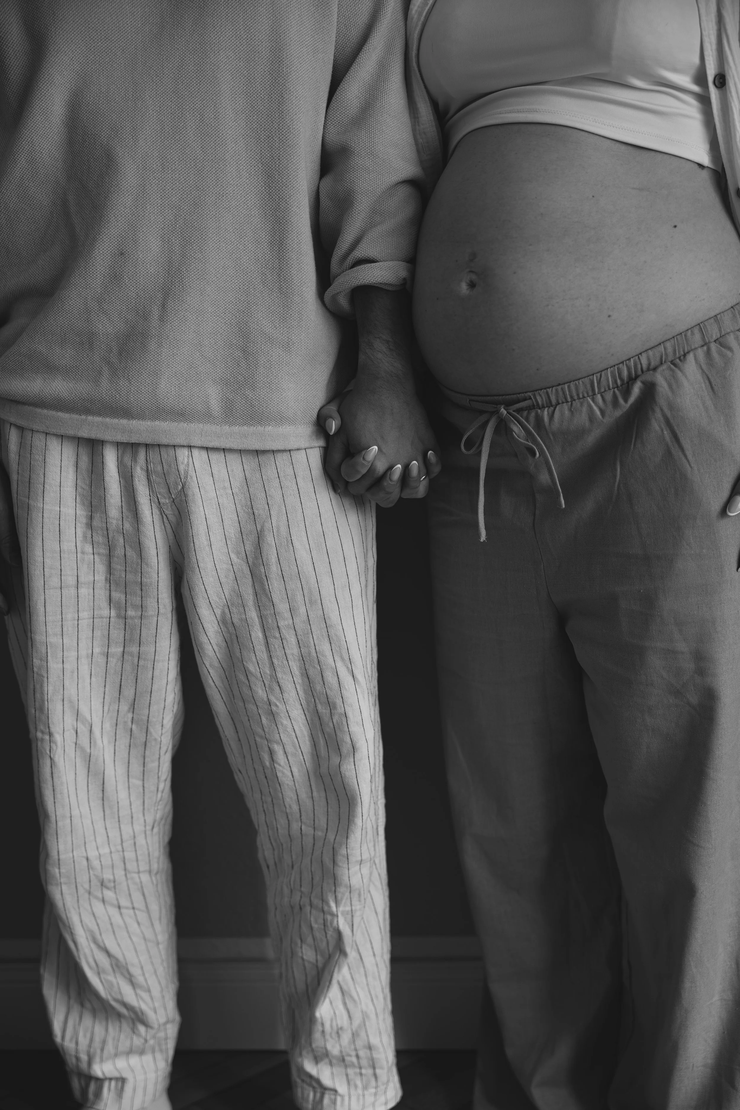 Ein Mann und eine schwangere Frau halten sich an den Händen. Die Frau trägt bequeme Jogginghosen, der Mann trägt gestreifte Hosen. Es ist eine Nahaufnahme, schwarz-weiß.