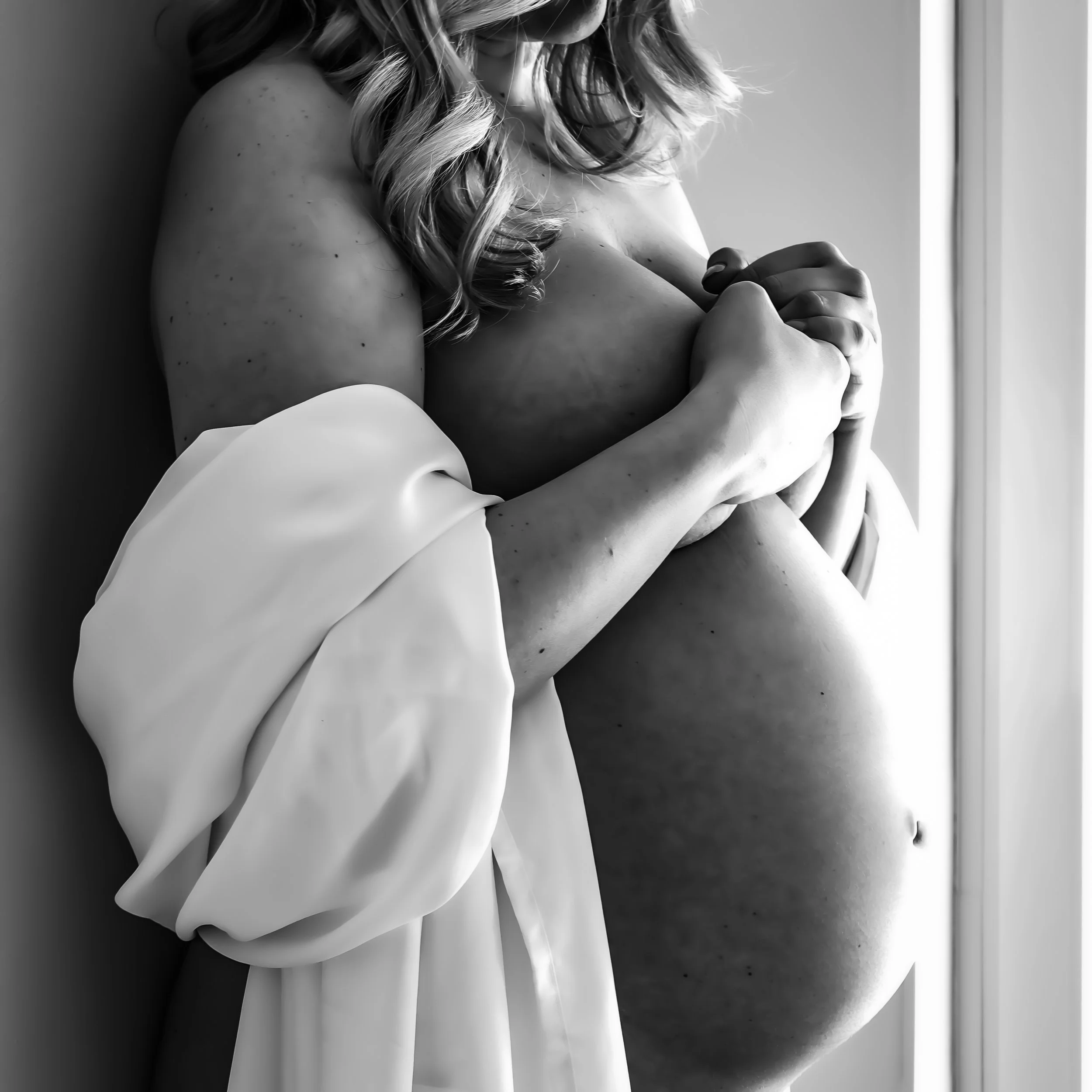 A black and white photograph of a woman with curled hair, partially dressed in a white fabric, holding her pregnant belly.